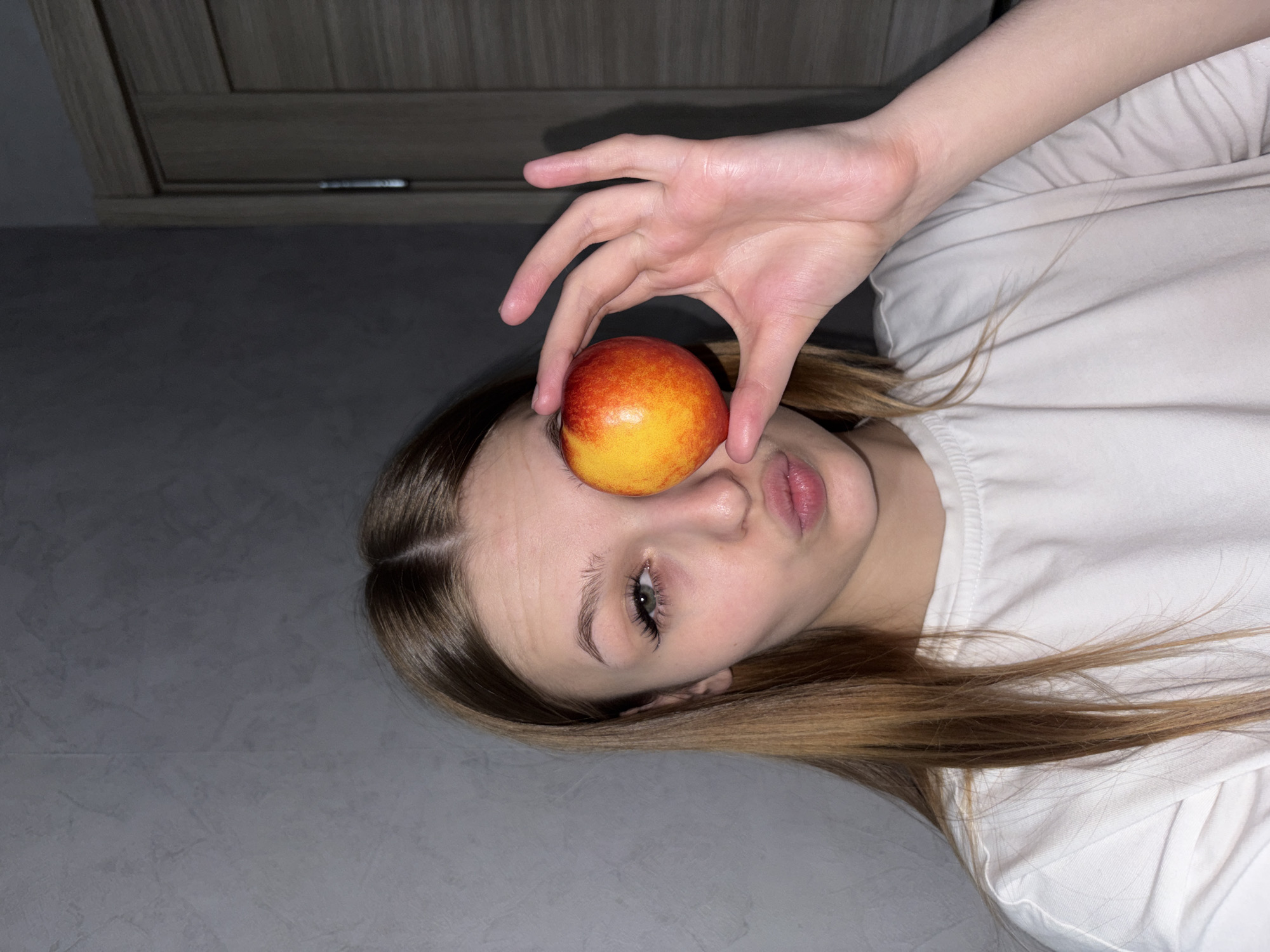 A young woman is lying down with a peach held over her left eye. She is wearing a white shirt. The background is a gray wall and floor, with a gray cabinet visible in the upper left corner. The setting appears to be indoors. No evidence suggests the location is Chaikovsky, Russia.