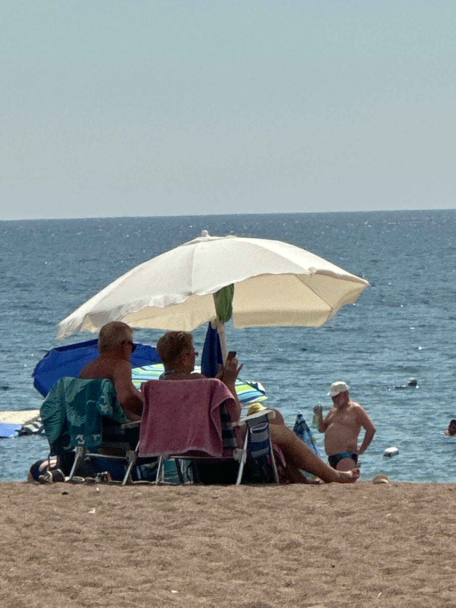 The image depicts a beach scene on a clear, sunny day. In the foreground, the sandy beach is visible with several beach chairs. One chair is draped with a teal towel, and another with a pink towel. Behind these chairs, two individuals are seated, facing away from the camera, under a large white umbrella. One person, on the left, has blonde hair and is wearing sunglasses. The other person, on the right, is wearing a pink towel over their shoulders and also appears to be wearing sunglasses. They are partially obscured by the umbrella.
In the middle ground, the calm, blue sea stretches to the horizon, meeting a pale blue sky. Several people are visible in the water. To the right of the umbrella, a man wearing a white cap and dark swim trunks stands in the water, holding a bottle. Further out in the water, other individuals are swimming or floating. The setting appears to be a beach resort, likely in the city of Boreti, Montenegro, given the context.