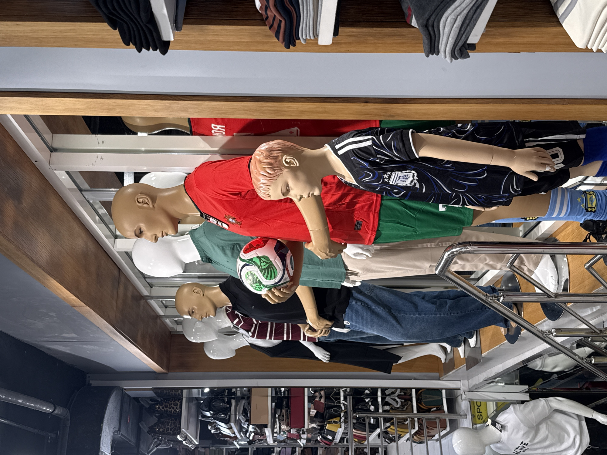 The image shows a row of mannequins dressed in sportswear displayed inside a clothing store. The mannequins are positioned on tiered platforms. From left to right, the first mannequin is a headless figure wearing a white t-shirt with the word "ACTIVE" printed on it. The next mannequin, also headless, is partially visible, wearing a dark top and blue jeans. The third mannequin is headless and wears a striped maroon and white shirt with a dark collar, paired with blue jeans. The fourth mannequin is headless and wears a black polo shirt with blue jeans. The fifth mannequin, a male adult, is wearing a green short-sleeved collared shirt and tan pants, and is holding a soccer ball. The sixth mannequin, a male adult, is wearing a red soccer jersey with a crest on the left chest and green shorts. A seventh mannequin, a young boy, is dressed in a black and white patterned soccer jersey and black shorts with white stripes. In the background, shelves are stocked with clothing and accessories. The store appears to be located in the city of Deira, United Arab Emirates, suggested by the presence of numerous retail shops and the general ambiance typical of the area.