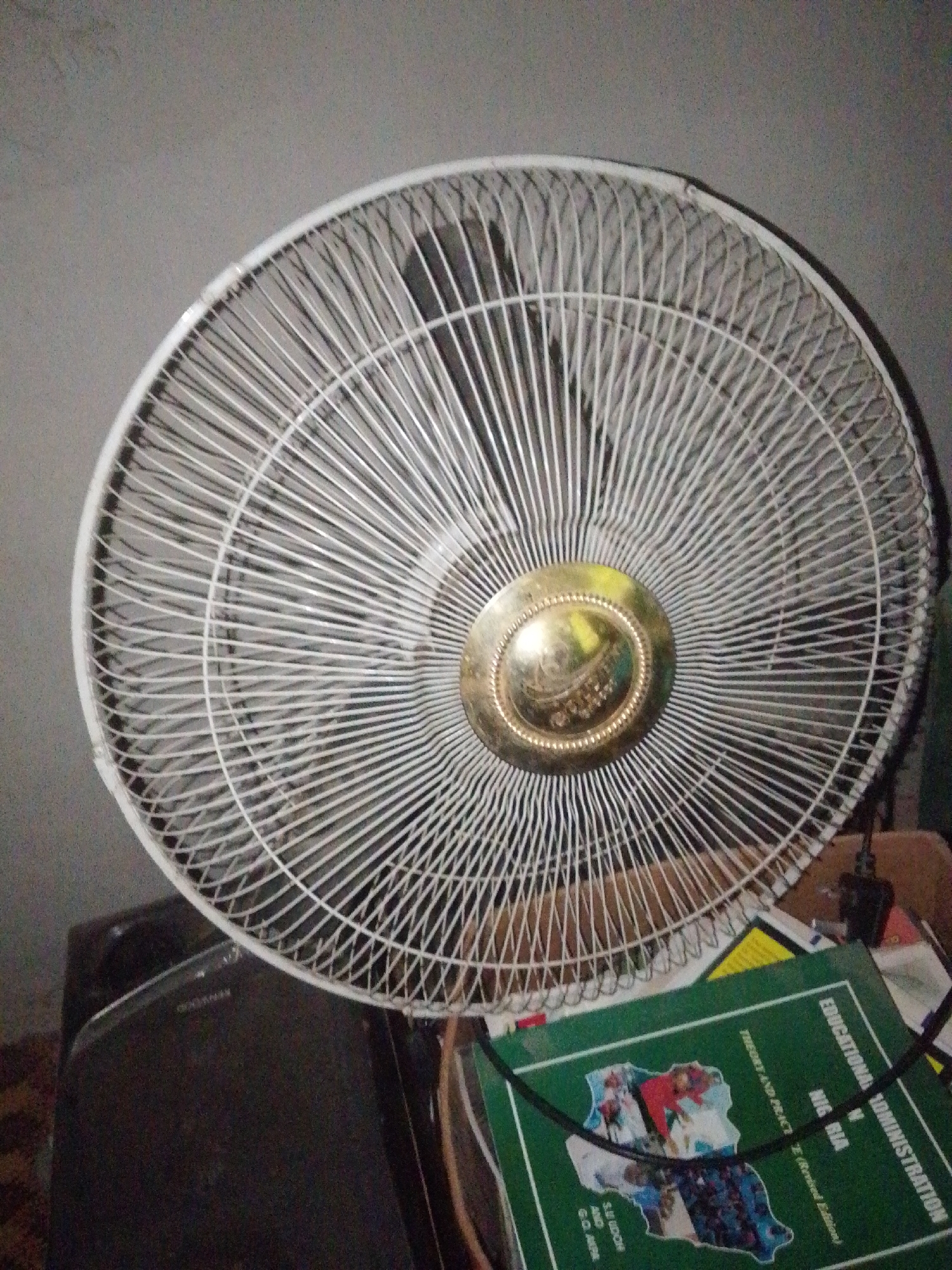 The image shows a close-up, indoor shot of a white oscillating fan. The fan is positioned in the foreground, taking up most of the frame. Its grille is intricate, with many thin, white wires forming a radial pattern. The central hub of the fan has a metallic, gold-colored emblem with some writing and a design, though the details are unclear. Behind the fan, a dark, rectangular object is partially visible, possibly a piece of furniture or electronics.
In the lower right portion of the image, a green book is laid out. The book has text in English, with prominent headings like "EDUCATIONAL ADMINISTRATION" and "NIGERIA." A map of Nigeria is printed on the cover, overlaid with various images and text, suggesting it is an educational textbook. The background is a plain, grey wall, and the lighting is dim, creating a slightly grainy texture across the image. The scene appears to be within a residential or office space. Given the presence of an educational textbook about Nigeria, this could be within a home, school, or office in Jalingo, Nigeria.