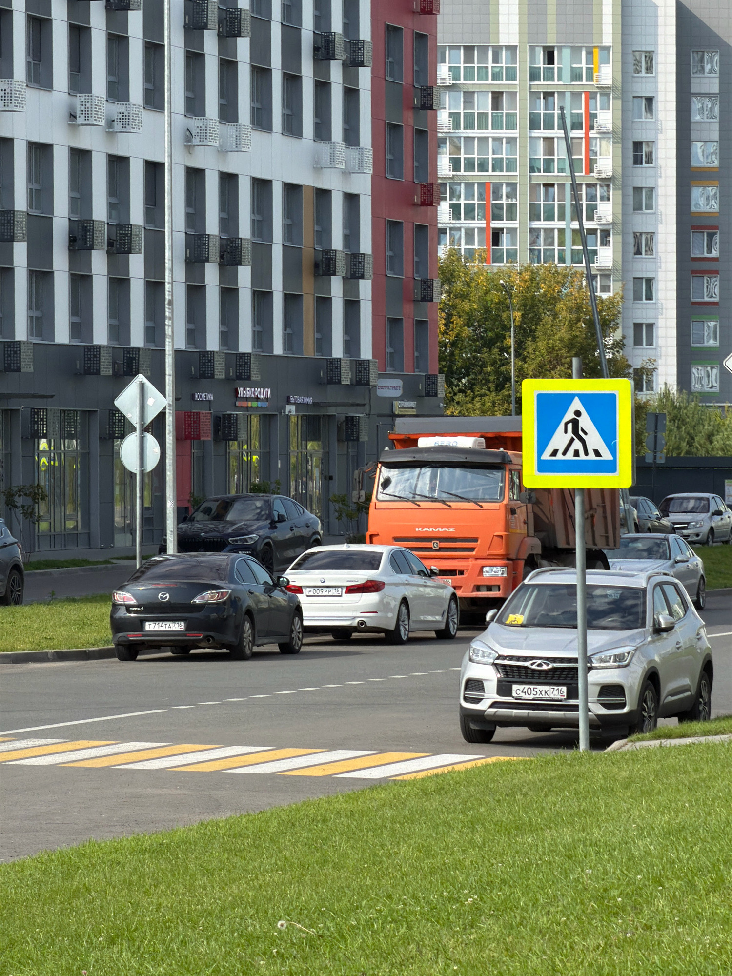 The image depicts a street scene in front of modern apartment buildings. In the foreground, a crosswalk is visible, with cars on the road. The cars include a dark-colored sedan, a white sedan, and a silver SUV. A large orange dump truck is parked further down the street, near a building with storefronts. A pedestrian crossing sign with a yellow border and a blue interior, featuring a silhouette of a person walking, is prominently displayed on a pole. The buildings in the background are tall and multi-story, with a mix of grey, white, and colorful facades, featuring numerous windows. Trees with green foliage are visible between the buildings and the road. The sky is overcast, suggesting daytime.