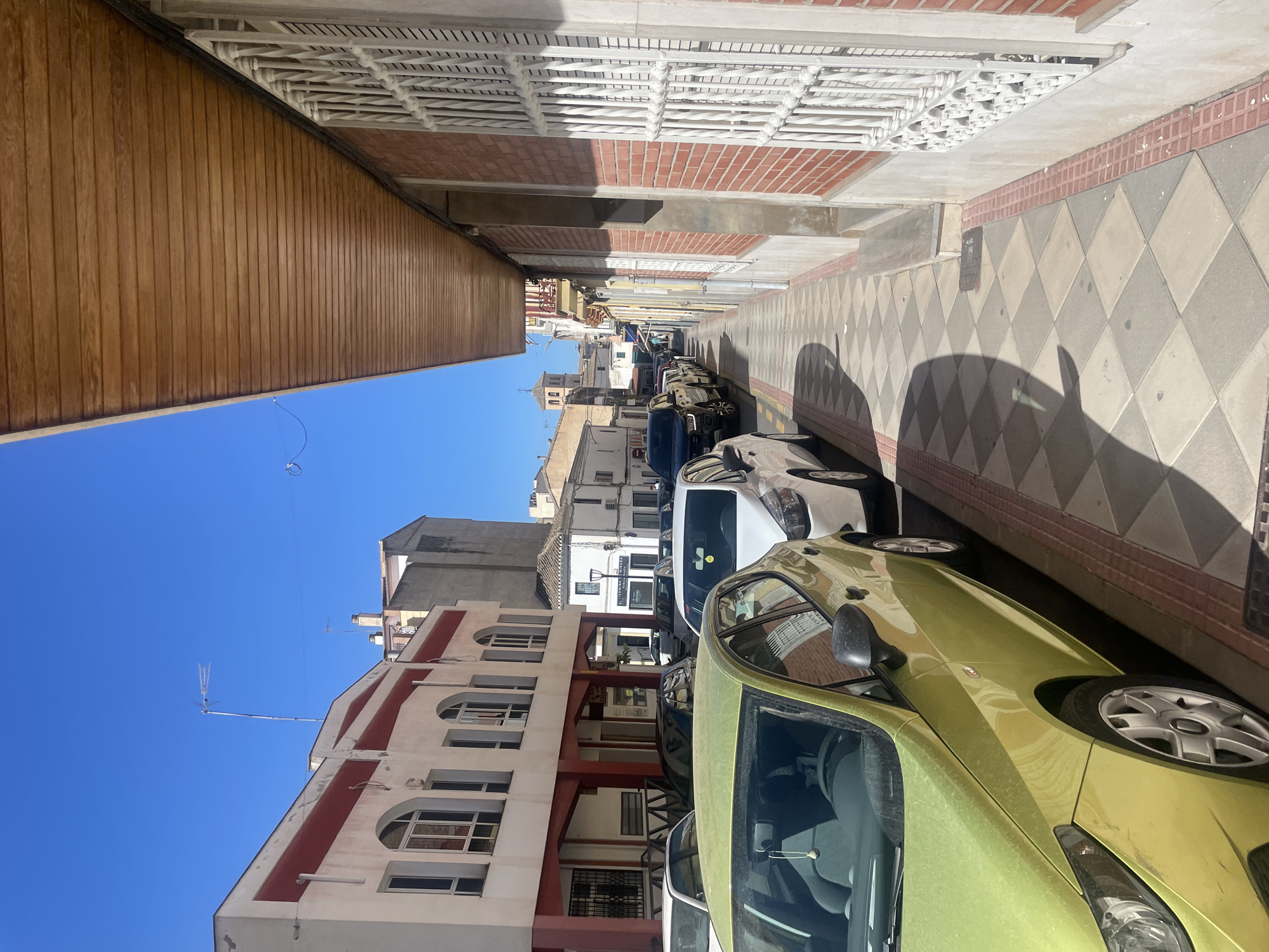 The image shows a street scene in Atarfe, Spain, on a bright, sunny day. The perspective is from ground level, looking down a sidewalk lined with parked cars. On the left, a light green car is prominently in the foreground, parked alongside other vehicles. Behind these cars, buildings with white facades and red accents are visible, featuring arched windows and awnings. The sidewalk itself is paved with a diamond pattern of beige and grey tiles, bordered by a red brick edge.
To the right, a building with a brick facade and a white metal grate covering a window runs along the sidewalk. Above this, a wooden soffit extends outwards, casting a shadow on the pavement. Further down the street, more buildings and parked cars are visible, receding into the distance under a clear blue sky. A distant church tower with a cross is visible in the background. No people are clearly depicted in the image.