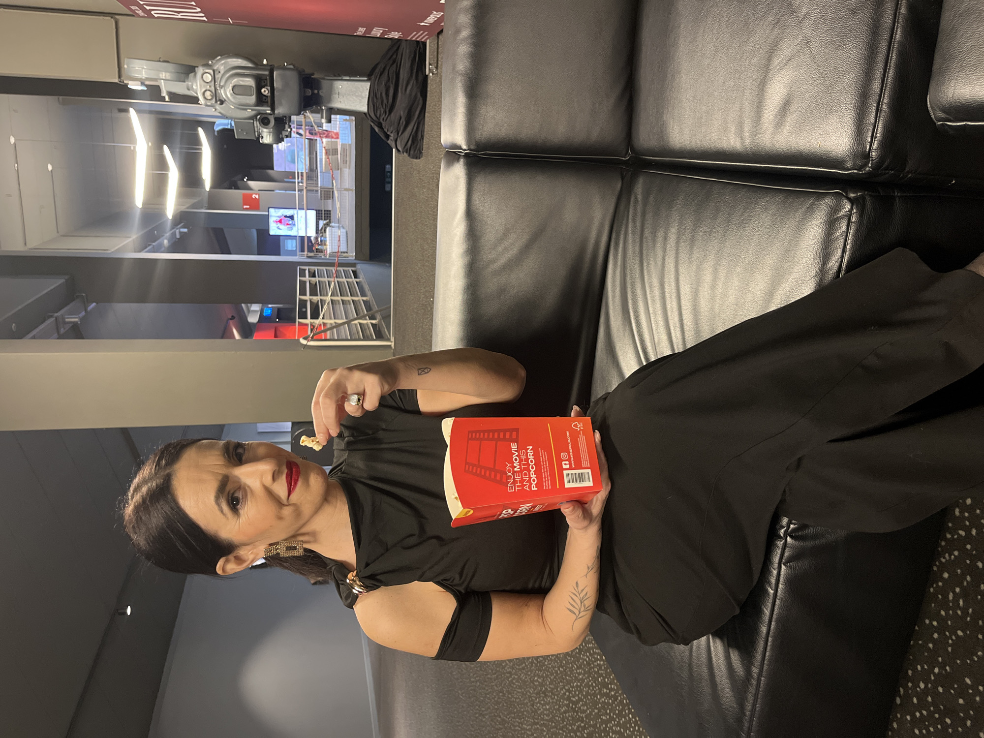 The image captures a woman seated on a black leather couch, appearing relaxed and enjoying a snack. She is dressed in black attire and holds a red box of popcorn, suggesting she may be at a movie theater or a lounge associated with one. The popcorn box reads "ENJOY THE MOVIE AND THIS POPCORN." She is holding a piece of popcorn in her hand, ready to eat. Her hair is dark, and she's wearing red lipstick and earrings. Tattoos are visible on her arms.
In the background, there is a glimpse of a well-lit interior space, potentially part of the same establishment. A camera rig or other professional equipment is visible, and it looks to be in the city of Luxembourg, Luxembourg.