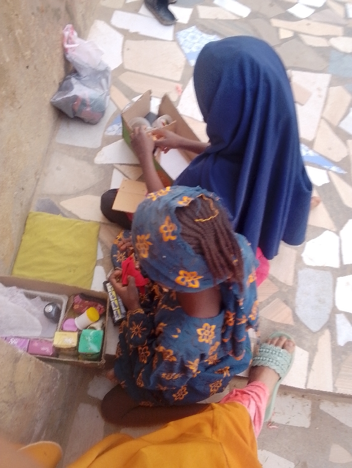 Two individuals are seated on a patterned tile floor. One, wearing a blue headscarf, faces away, working with items in a cardboard box. The other, donning a floral blue headscarf and dress, holds a red item, also near a box filled with soap and other toiletries. A plastic bag of items sits nearby, and a yellow cloth pad is on the floor next to the person in front. A pair of feet is wearing a sandal on the bottom-right.