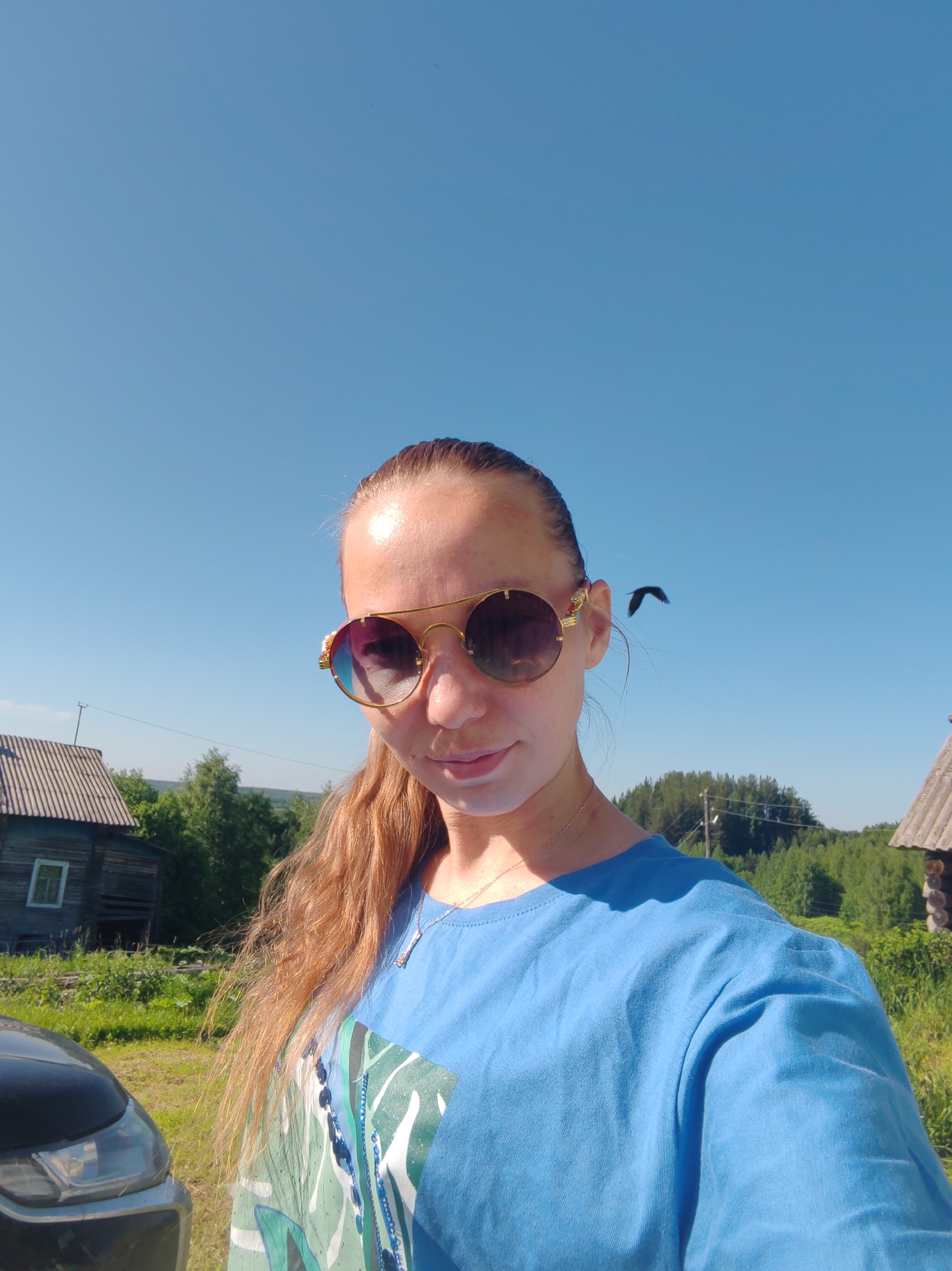 A selfie captures a young woman standing outdoors on a bright, sunny day. She is wearing a blue t-shirt with a green and white abstract design and a delicate gold chain necklace. Her long, auburn hair is pulled back into a ponytail, and she is sporting large, round, gold-rimmed sunglasses with colorful lenses. Her face is tilted slightly upwards. In the background, a wooden house with a corrugated roof stands to the left, and another wooden structure is visible to the right. A lush green landscape with trees and grass surrounds the buildings. The sky is a clear, vibrant blue, with a single dark bird in flight to the right of the woman's head. Part of a dark-colored car is visible in the lower left corner of the frame. The setting appears to be a rural area, potentially in the city of Chuchlem, Russia, given the architecture and vegetation.