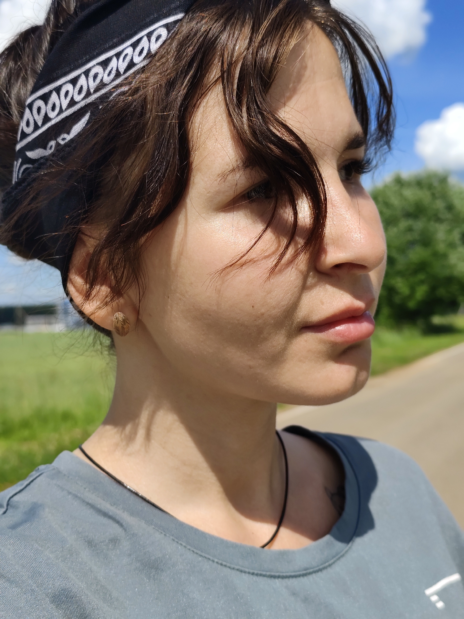 In the city of Foky, Russia, a young woman with dark, curly hair is pictured in a medium close-up, facing to the right. She is wearing a black bandana with a white paisley pattern around her head and a grey t-shirt. A thin black necklace is visible around her neck, and a small, patterned stud earring is in her right ear. Her left cheek shows a subtle shadow from the bandana, and a few strands of hair fall across her forehead and cheek. Her skin is illuminated by sunlight, creating a warm glow. In the background, a blurred green landscape is visible, with a paved road and a bright blue sky with white clouds. The woman's facial expression is neutral, and her gaze is directed away from the camera.
