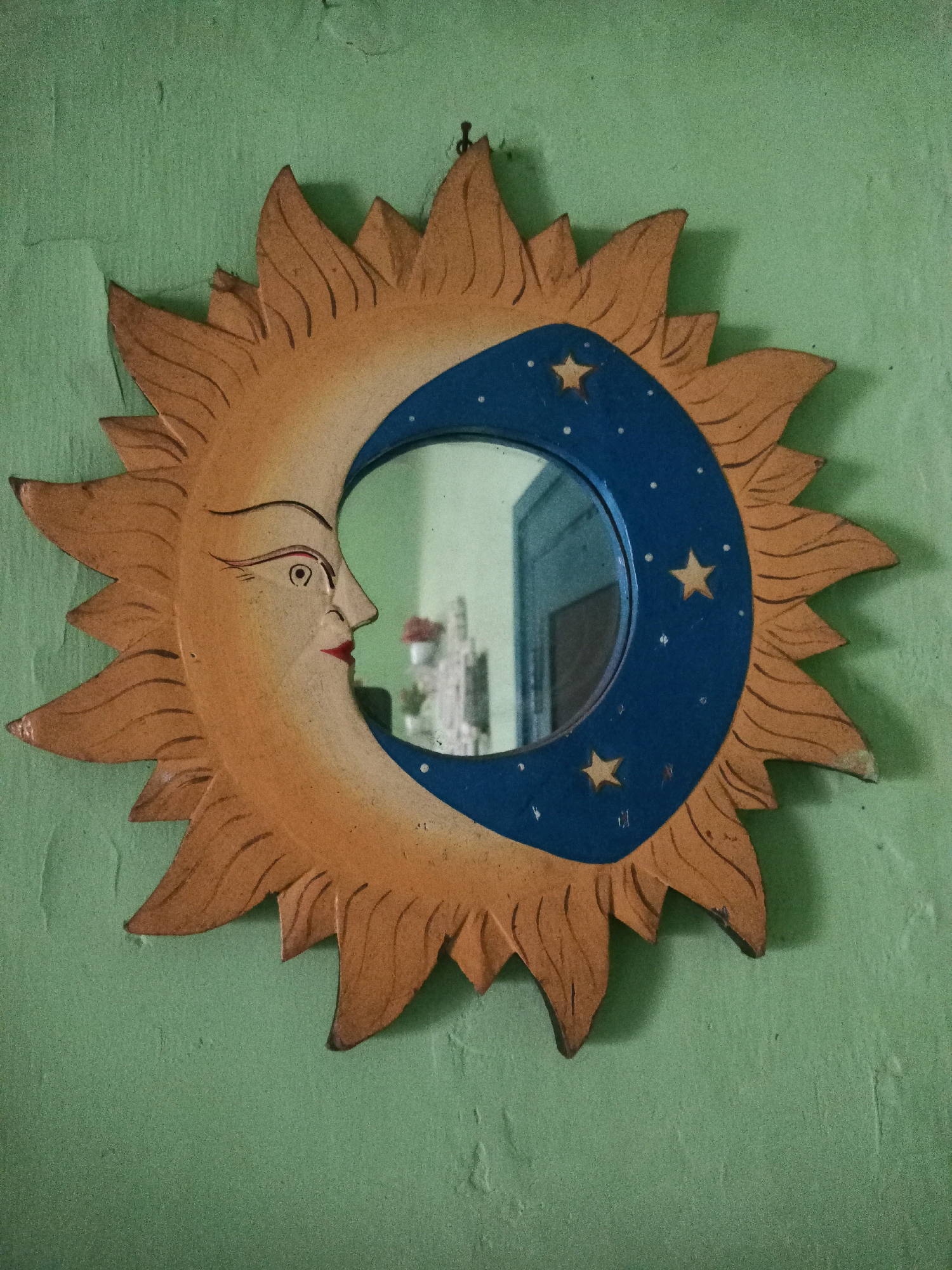 This is a close-up, eye-level, static shot of a decorative mirror hanging on a light green, textured wall. The mirror is shaped like a sun with a crescent moon face. The sun's rays are depicted as pointed, orange-brown wooden cutouts radiating from the central mirror. The crescent moon, which forms the frame of the actual reflective surface, is painted blue and decorated with small white dots and three yellow stars. The moon's face, carved into the wooden sun frame, is in profile and features painted details: an arched eyebrow, an eye with a pupil, a pointed nose, and red lips. The background wall appears to be a plain interior wall, possibly in a residential setting. No people are visible in the image. The context of Malang, Indonesia, is not visually apparent from the image itself.