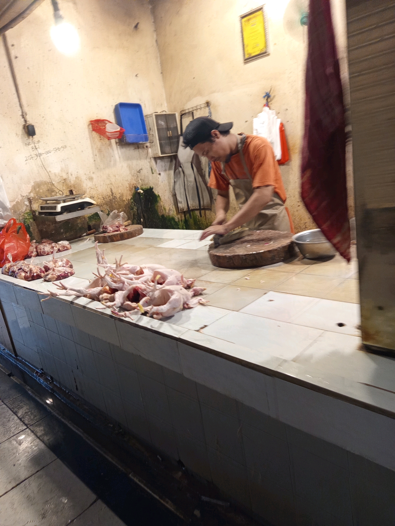 In a butcher stall located in the city of Karawang, Indonesia, a man wearing an orange shirt and a beige apron is actively engaged in butchering poultry. He is positioned behind a large, round wooden cutting board, meticulously preparing raw chickens, which are spread out on the tiled counter in front of him. To the left of the butcher, there are packaged cuts of meat and red plastic bags, suggesting a busy marketplace environment. The stall itself is utilitarian, with a tiled counter and walls, and overhead lighting illuminating the workspace. In the background, various objects are visible, including a weighing scale, a blue plastic bin, and a framed certificate hanging on the wall. The scene captures a typical moment of commerce and food preparation within a local Indonesian market.