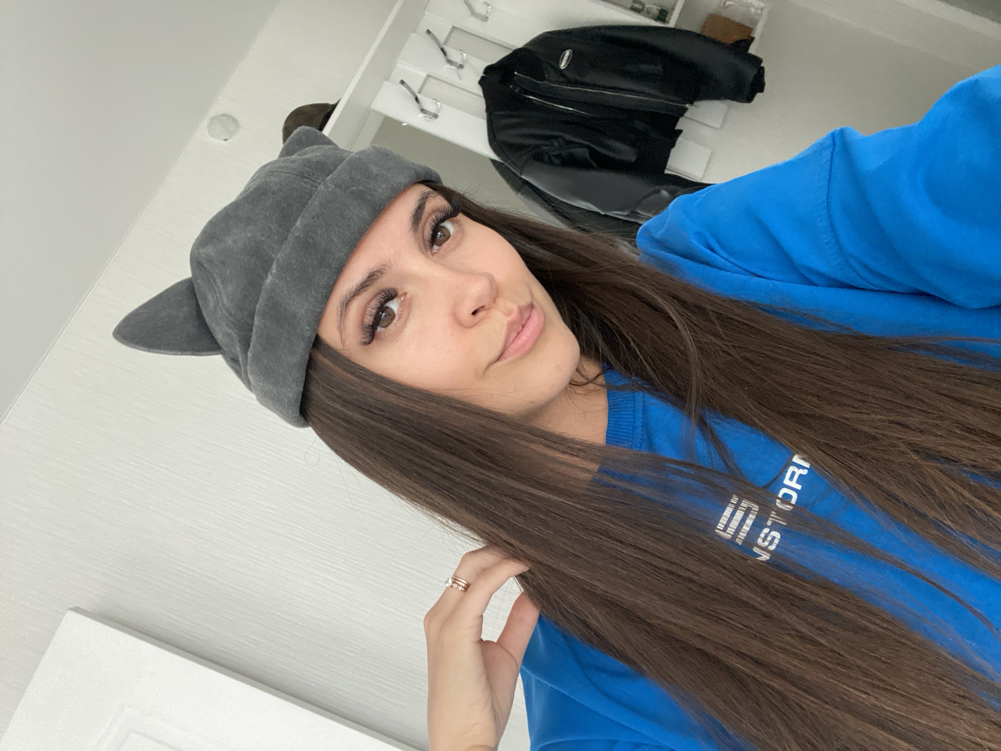 The image is a selfie taken indoors. A young woman with long, brown hair is wearing a grey beanie with cat ears and a bright blue sweatshirt with white lettering on it. Her left hand is resting on her hair, and she is wearing a ring on her ring finger. In the background, there is a white wall with a door on the left side and a closet or coat rack with a black jacket hanging on it to the right. The lighting suggests it is daytime. There is no information within the image to indicate the location is in Chaikovsky, Russia.