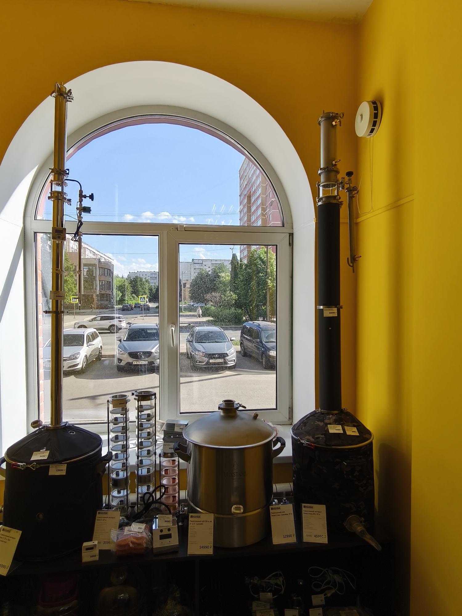 The image shows an indoor view from a shop, looking out through a large arched window. The shop has bright yellow walls. Inside the shop, on a dark shelf in the foreground, are several items including large metal pots, clear cylindrical containers with small items inside, and smaller electronic devices. Price tags are visible on some of these items. Two tall, thin distillation apparatuses, one on the left and one on the right, are positioned in front of the window. The one on the left is primarily gold-colored metal, while the one on the right is mostly black with some metal components.
Through the window, a street scene is visible. Several cars are parked on the street and on the side of the road. Buildings, including a multi-story brick building, are present across the street, along with trees and a clear blue sky with some clouds. Yellow pedestrian crossing signs are visible on the street. The exterior of the buildings appears to be in Yoshkar-Ola, Russia, based on the visible text on the price tags which includes Russian currency symbols.