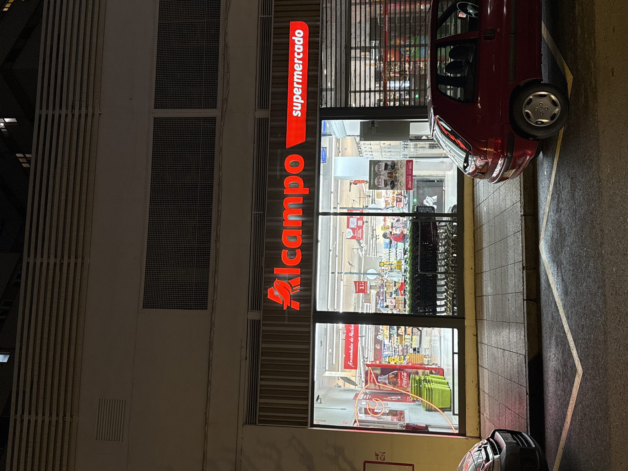 The image shows the exterior of an Alcampo supermarket at night in Oviedo, Spain. The illuminated "Alcampo Supermercado" sign is vertically aligned along the building's facade. Inside, the supermarket is brightly lit, revealing aisles of products, shelves, and promotional displays. Shopping carts are visible near the entrance. A red compact car is parked partially blocking the entrance, with its rear end angled towards the building. A second vehicle is partially visible at the bottom edge of the picture. The building's facade consists of gray concrete walls with rectangular grilles and vertical vents. The sidewalk next to the building features a painted yellow line and a dark grey asphalt surface.