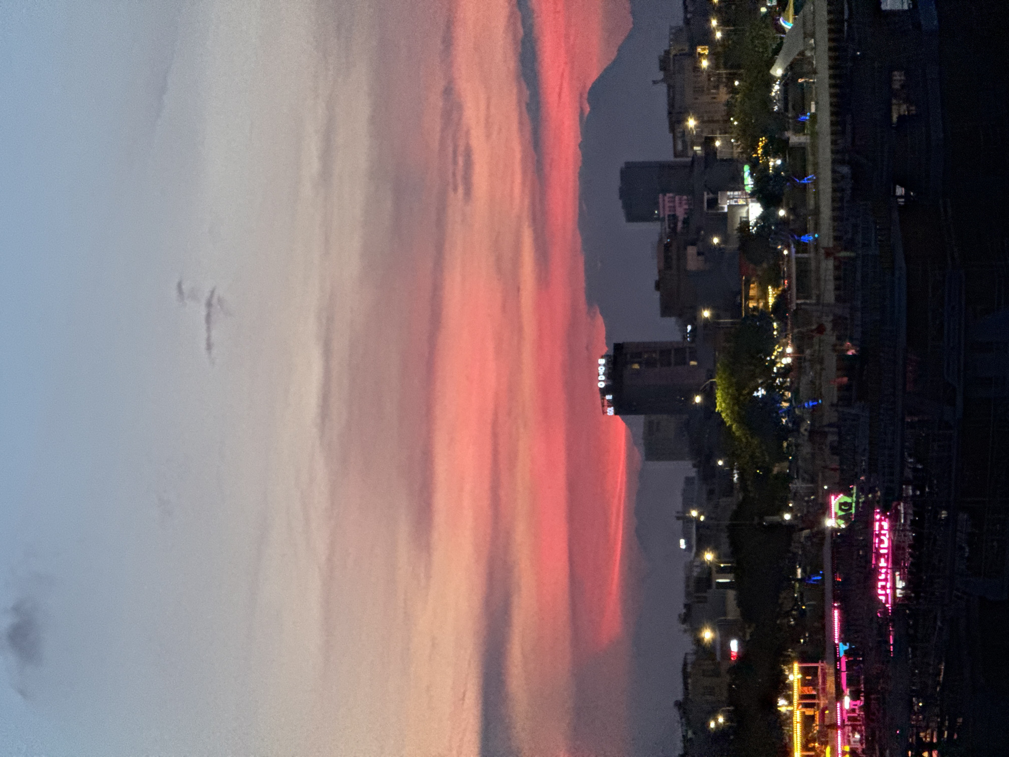 This is a vertical, wide-angle, dusk shot of a city skyline in Da Nang, Vietnam. The left half of the image is dominated by a gradient sky, transitioning from a pale blue at the top to soft pinks and oranges towards the center, suggesting sunset or twilight. Faint clouds are visible in the sky. The right half of the image shows a city scene, with buildings of varying heights illuminated by streetlights and colorful neon signs. Trees are interspersed throughout the urban landscape. The overall mood is one of urban vibrancy against a dramatic natural sky.
The composition places the vast expanse of the sky on the left and the bustling city on the right. The buildings are densely packed, with many windows reflecting light. Several of the neon signs are brightly colored, featuring pinks, yellows, and blues, adding a dynamic and festive atmosphere to the scene. The lights create a sense of activity and life in the city at dusk.
Notable details include the subtle texture of the clouds in the sky and the sharp definition of the illuminated city elements. The time of day is clearly indicated by the twilight sky and the extensive artificial lighting. The presence of multiple stories of buildings, streetlights, and the types of neon signage are strong cues for an urban setting, specifically a commercial or entertainment district within a city.
The image is rich in color contrast, with the soft, natural hues of the sky juxtaposed against the bold, artificial lights of the city. This creates a visually striking effect, capturing the transition from day to night in a dynamic urban environment.
No people are clearly discernible as subjects in this image. The focus is on the interplay between the natural sky and the man-made city. The overall impression is one of urban beauty and the lively energy of a city at twilight.