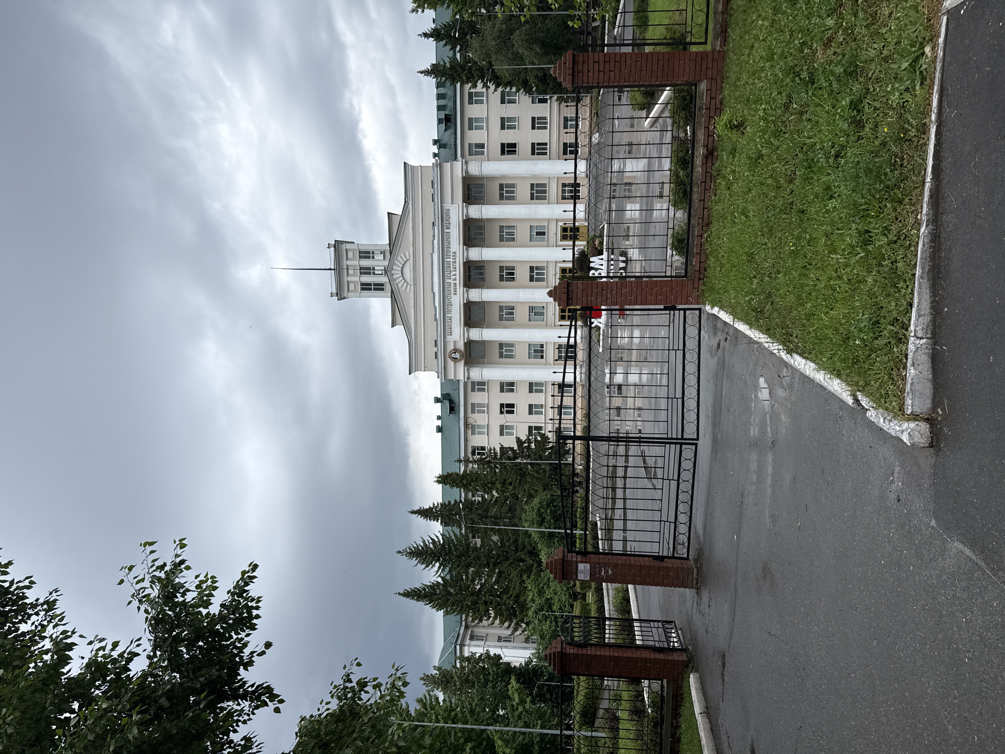 This media file shows an exterior view of a large, imposing building with classical architectural elements, likely an educational institution. The building is light-colored, with a prominent central section featuring a colonnade of tall, white columns and a stepped roofline. A tower-like structure rises from the center of the building. In front of the building, there is a black metal gate, supported by brick pillars, and a paved pathway leading to it. To the right of the pathway is a well-maintained patch of green grass. Lush green trees, including several tall coniferous trees, are visible around and in front of the building. The sky is overcast and a soft grey, suggesting it might be daytime, possibly late afternoon or early morning, with a light cloud cover. The overall scene appears calm and somewhat somber due to the weather.
Visible text on the building reads: "КАЗАНСКАЯ ГОСУДАРСТВЕННАЯ АКАДЕМИЯ ВЕТЕРИНАРНОЙ МЕДИЦИНЫ ИМ. Н.Э. БАУМАНА". This translates to "Kazan State Academy of Veterinary Medicine named after N.E. Bauman". This text clearly indicates the location and nature of the building. The presence of a fence and gate suggests it is a formal entrance to the institution's grounds.
