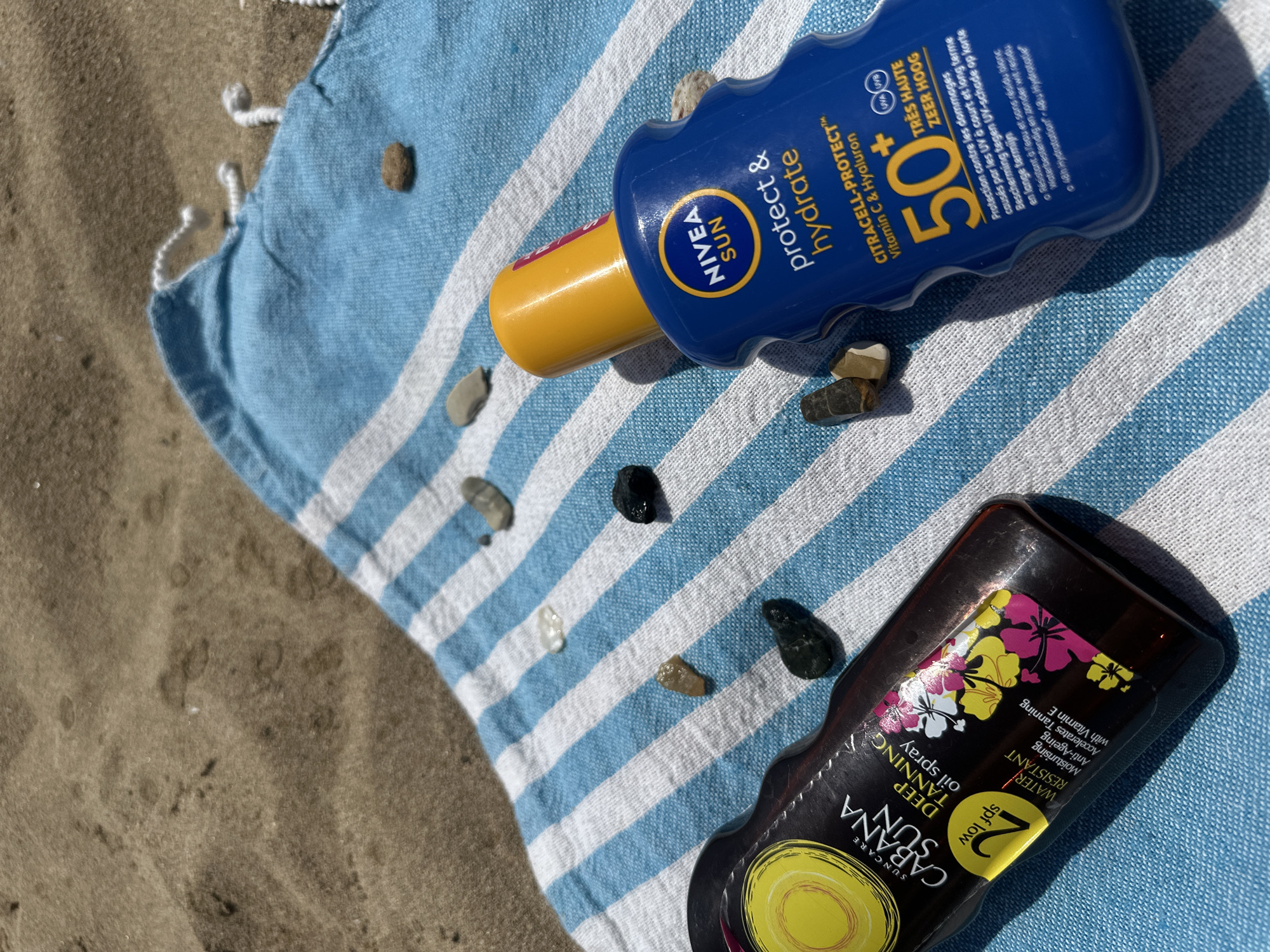 The image shows a beach towel with blue and white stripes on a sandy surface. Two bottles of sunscreen are placed on the towel, along with several small pebbles. On the left is a bottle of Suncare Cabana Sun Deep Tanning Oil Spray with SPF 2. On the right is a blue bottle of Nivea Sun Protect & Hydrate with SPF 50. The scene is illuminated by direct sunlight, casting shadows on the towel and sand, suggesting an outdoor environment. The location of Peyia, Cyprus is not visually represented in the image itself; this information is provided externally. There are no people or actions depicted.