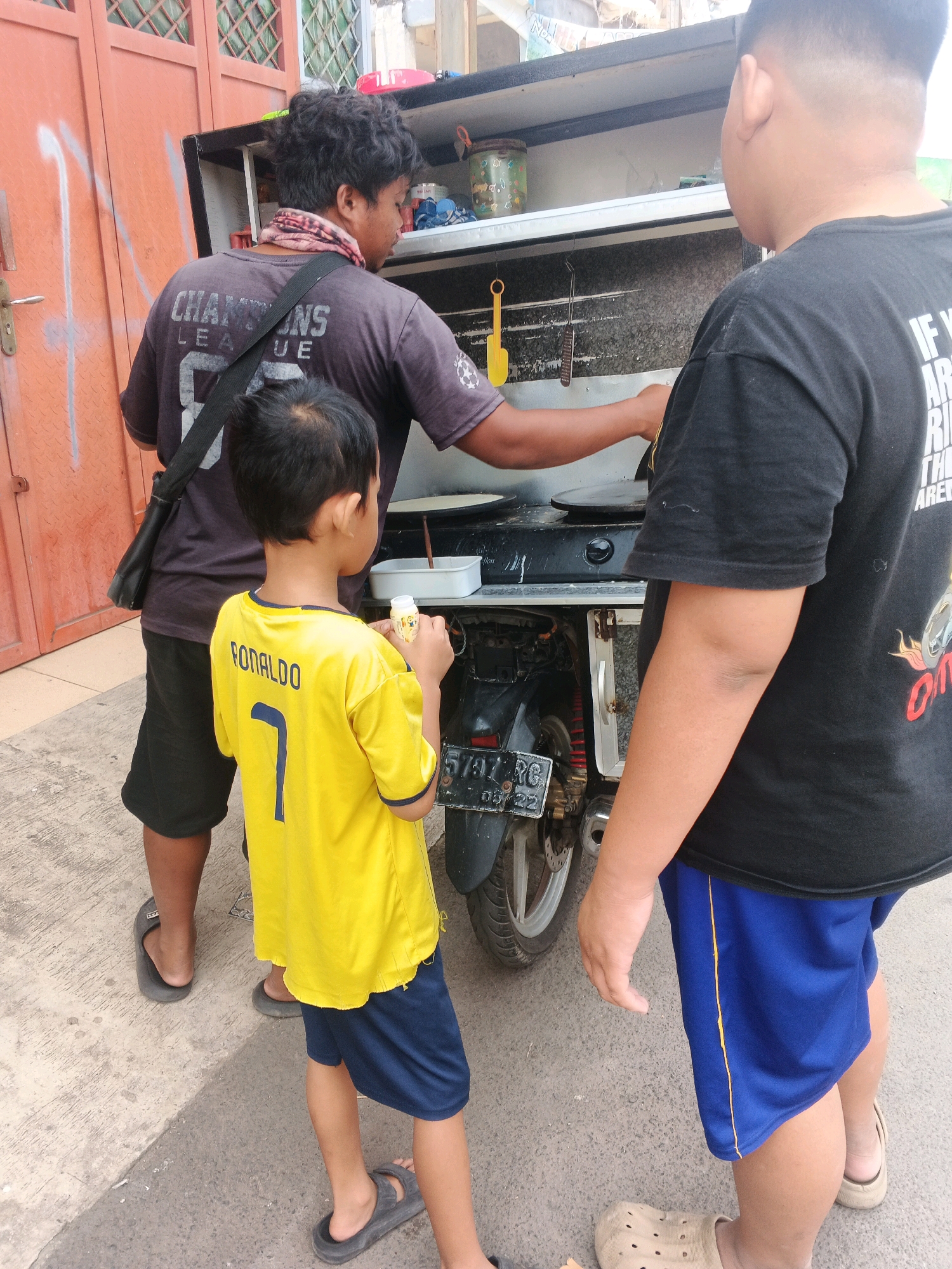 A street vendor, seen from behind, stands at a food cart, cooking. He wears a dark t-shirt with "CHAMPIONS LEAGUE" printed on it and a patterned neck gaiter. A young boy, also viewed from behind, stands to his left, wearing a yellow soccer jersey with "RONALDO 7" on the back and blue shorts. To the vendor's right, another person, a child or young adult, stands with their back to the camera, wearing a black t-shirt and blue shorts. The food cart appears to be motorized, possibly a repurposed motorcycle, and is parked on a paved street. In the background, there's an orange door with a diamond pattern, and other structures suggesting an urban or semi-urban setting. The image captures a moment in a public space, likely a street or market in Kadu, Indonesia, where food is being prepared and sold.