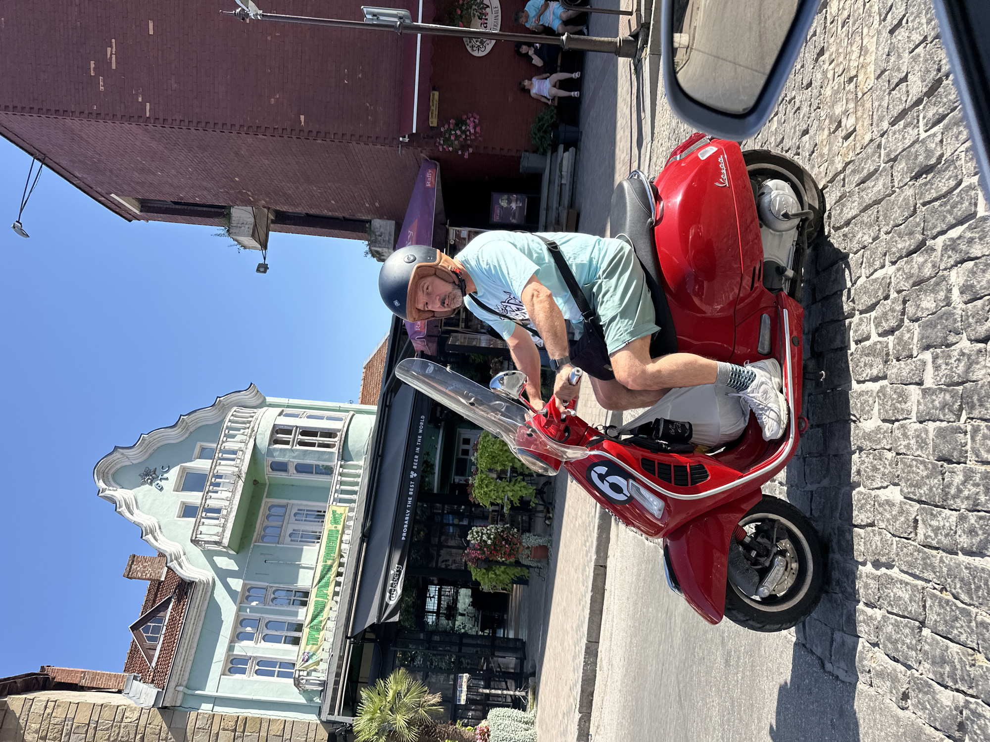 A man with a beard and a helmet rides a red Vespa scooter down a cobblestone street in Veliko Tarnovo, Bulgaria. He is wearing a light blue t-shirt and light green shorts. In the background, there is a building with a mint green facade, a balcony, and a banner that reads "GTC". To the right of the mint green building, there is a red brick building with a sign that says "Raffy". Several people are seated on a bench outside the red brick building. The sky is clear and blue.