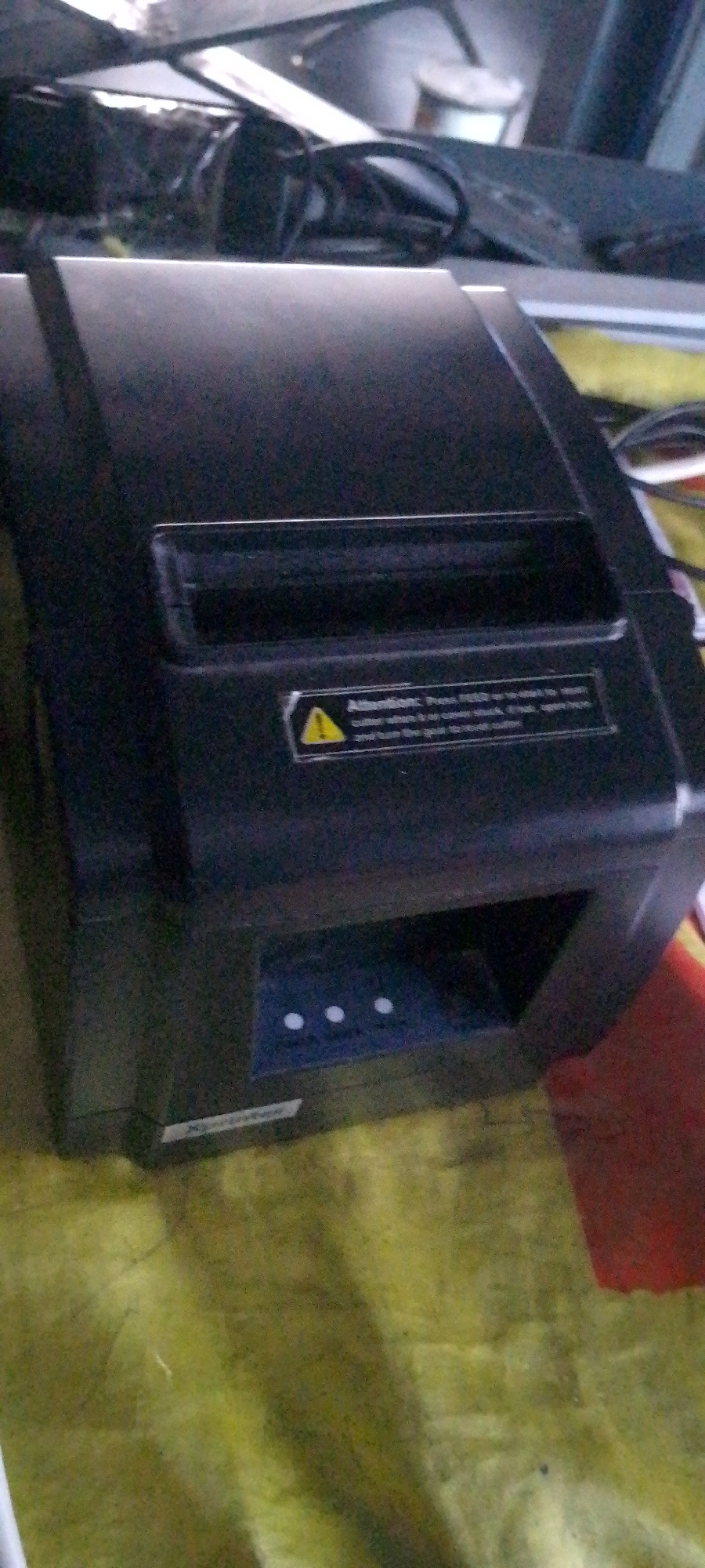 The image shows a black thermal printer on a green and yellow patterned surface. The printer has a slot at the top for paper to feed through, and a label with a yellow warning triangle and text is visible on its front. Below the paper slot, there are three small circular buttons. The background is out of focus, but appears to be a cluttered workspace with some metallic objects. There are no people present in the image. The lighting is dim, and the image has a grainy quality. The location is Unknown, Unknown.