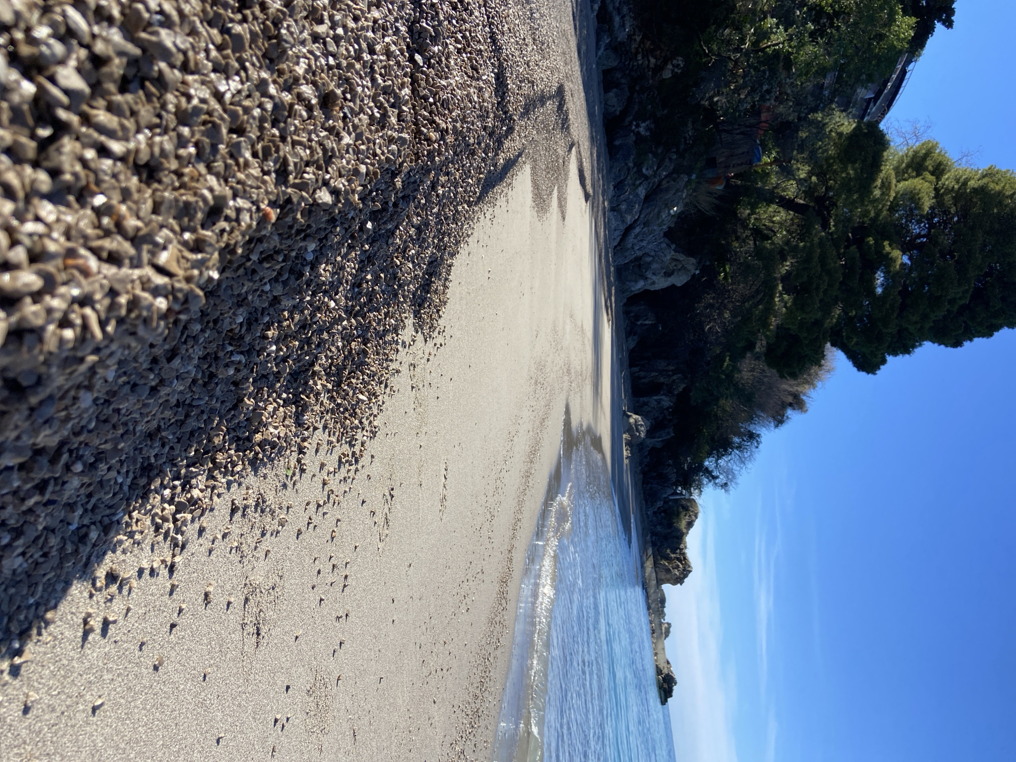 The image depicts a beach in Budva, Montenegro. The foreground shows a section of coarse gravel transitioning to sandy shore near the water's edge. The water is clear and calm. Vegetation and rocky formations are visible along the right edge of the frame, partially obscuring structures atop the rocks. The sky is clear and blue. The perspective is canted slightly.