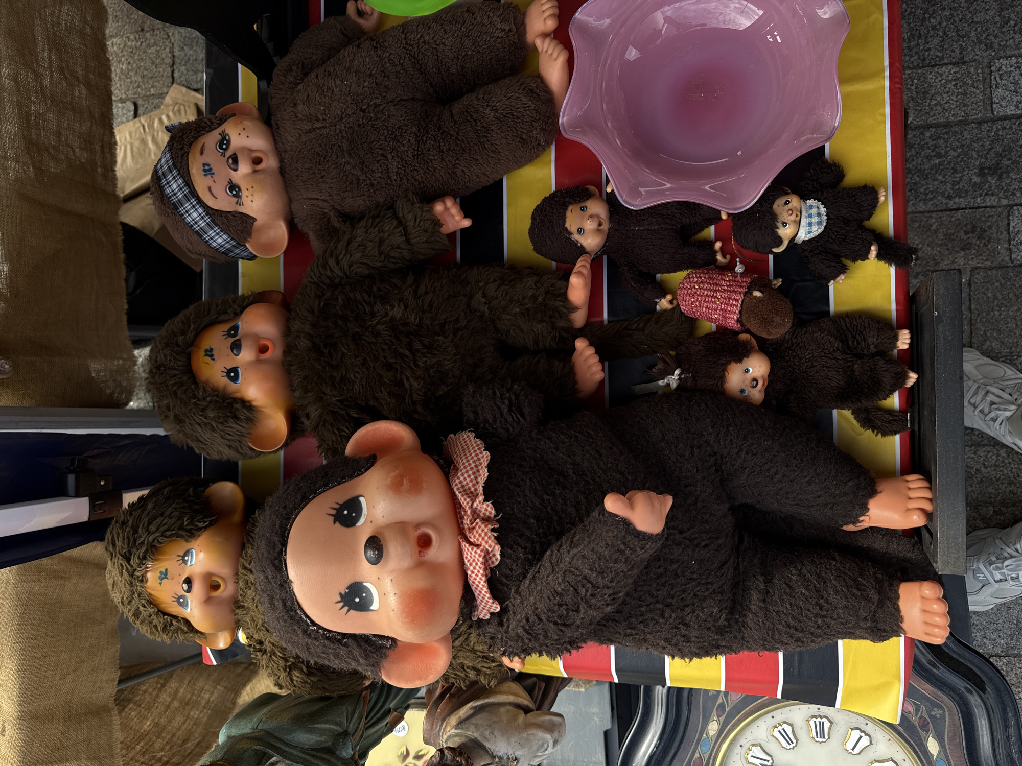 This is a high-angle, eye-level shot of a collection of Monchhichi dolls and other toys displayed on a market stall. The dolls are all made of brown fur with plastic faces and hands. The largest doll is in the foreground, wearing a red and white checkered collar. Behind it, several other larger Monchhichi dolls are visible, some with patterned headbands. Smaller Monchhichi dolls are also present, as well as a pink glass bowl, and a vintage clock face. The stall appears to be set up outdoors, with a rough-textured fabric canopy overhead and a paved ground surface. The overall impression is of a quirky, vintage toy stall.