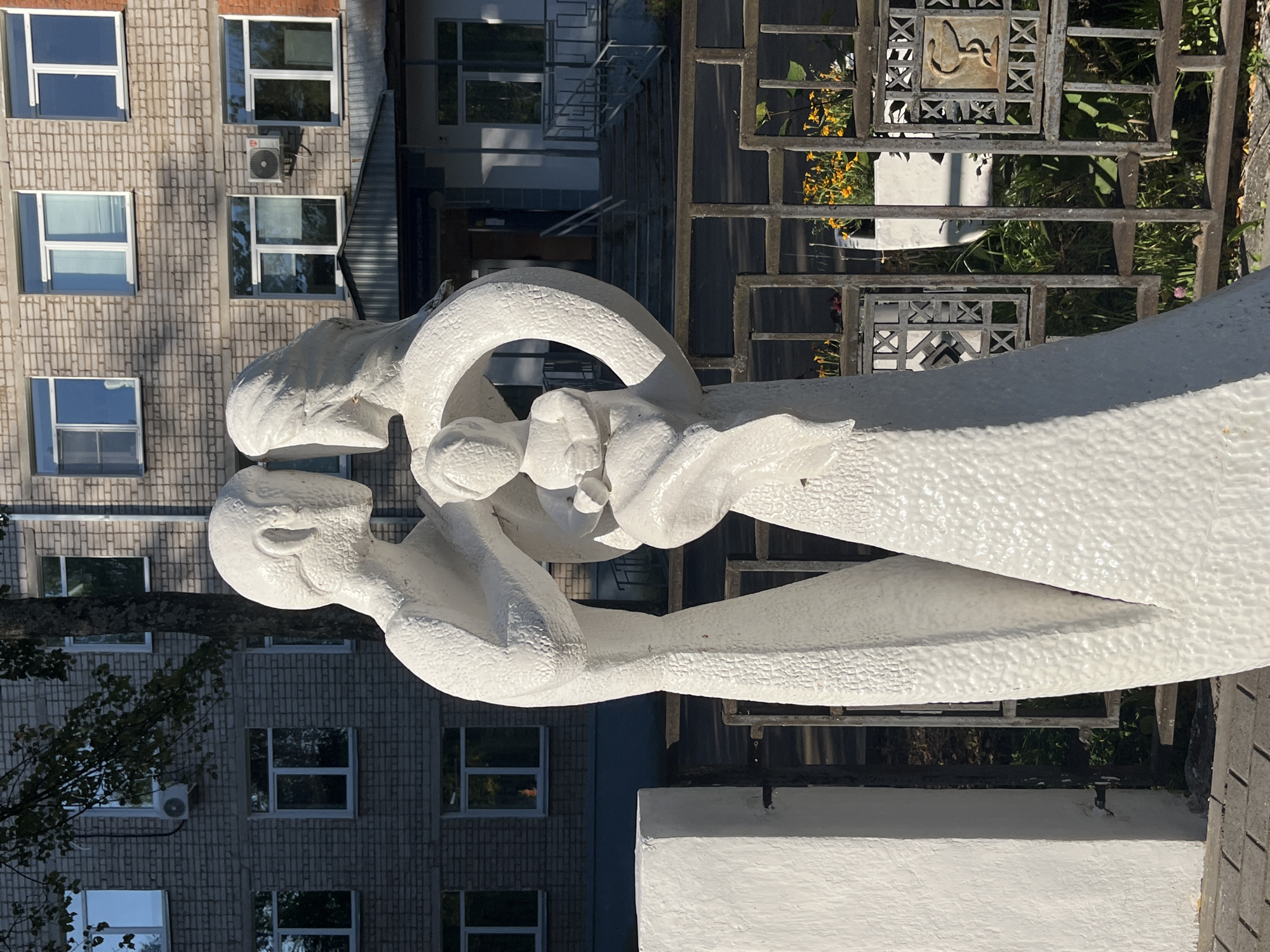 A white, textured sculpture of a family stands in front of a brick building. The sculpture depicts two adult figures, likely parents, embracing and holding a child between them. The adult figures are stylized, with smooth, rounded heads and bodies. The building behind them has multiple windows, some of which are open, and an air conditioning unit is visible on one of the walls. A metal fence with decorative elements runs in front of the building. Behind the fence, there are some plants and flowers. The ground in the foreground appears to be paved with interlocking bricks. The overall scene suggests an outdoor public space, possibly a park or the grounds of an institution.
This sculpture is located in the city of Chaikovsky, Russia, at the entrance to the Chaikovsky Children's Hospital.