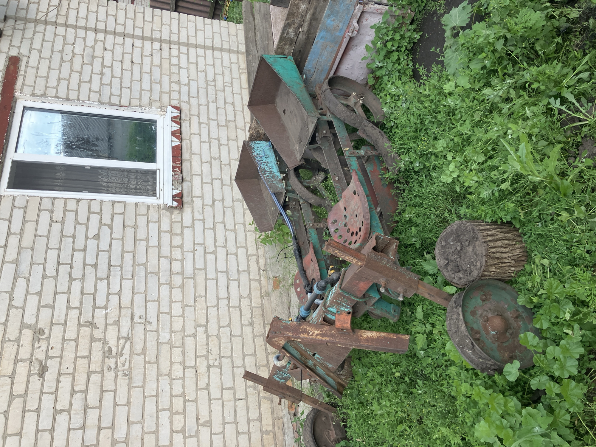 An old, rusty farm planter sits in overgrown grass in front of a white brick building with a single window. The planter has two metal hoppers, a seat, and wheels. To the right of the planter is a short, rough-hewn wooden log. The building appears to be a shed or an outbuilding. The overall scene suggests a rural or semi-rural setting. The image is taken outdoors during the day.