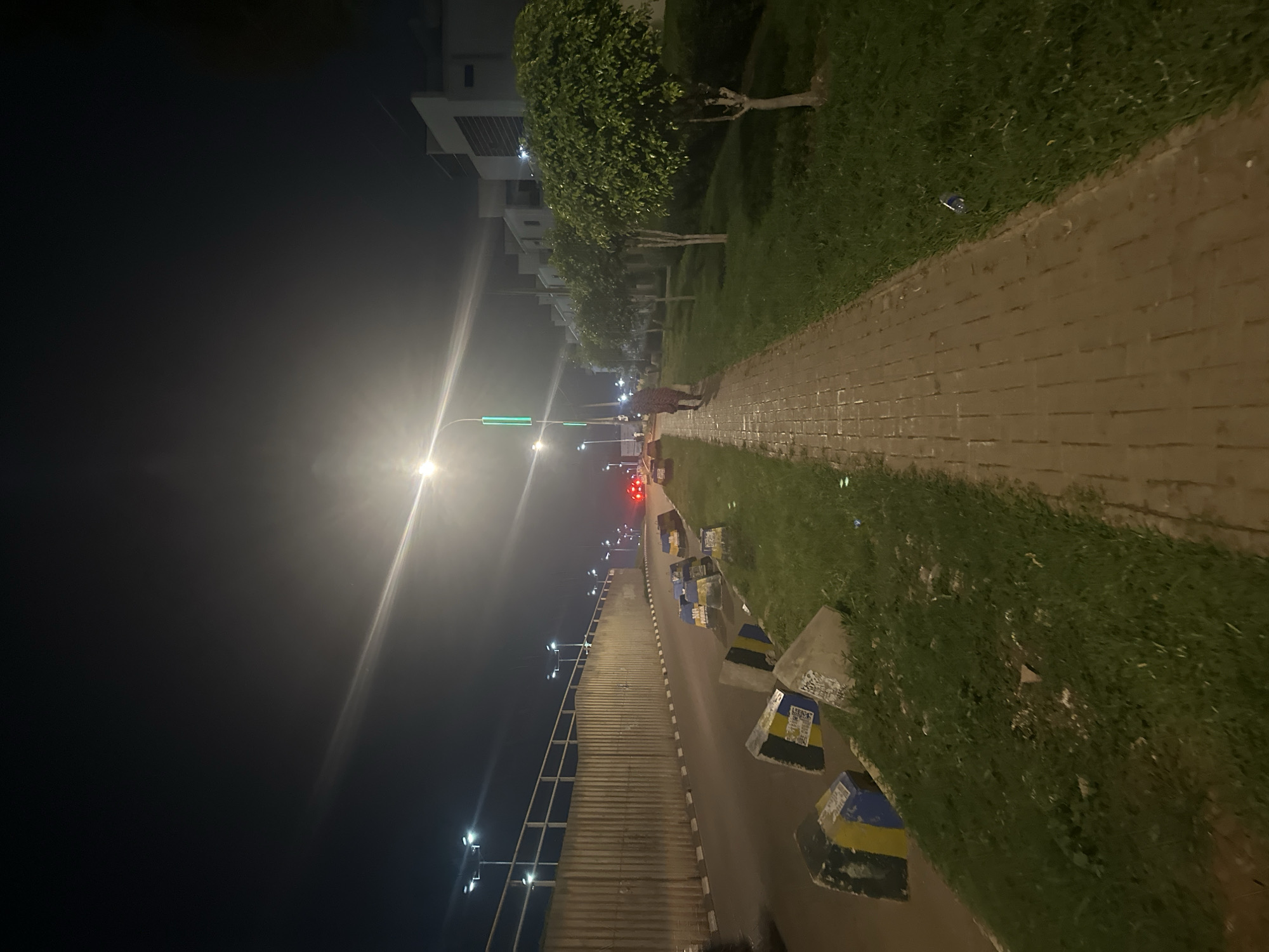 The image captures a nighttime scene along a street in Abuja, Nigeria. A paved pedestrian walkway, illuminated by streetlights, runs parallel to a road bordered by a concrete barrier and grassy areas. Several large concrete blocks, painted with yellow and blue stripes, are scattered along the edge of the road and grass, possibly serving as traffic barriers or decorative elements. Across the road, modern residential buildings with white facades and dark window accents are visible. A single pedestrian, dressed in dark clothing, walks away from the camera on the sidewalk. The sky is dark, with a bright light source causing a lens flare effect, likely from a streetlight. The environment appears to be an urban or suburban setting, with manicured trees lining the walkway.