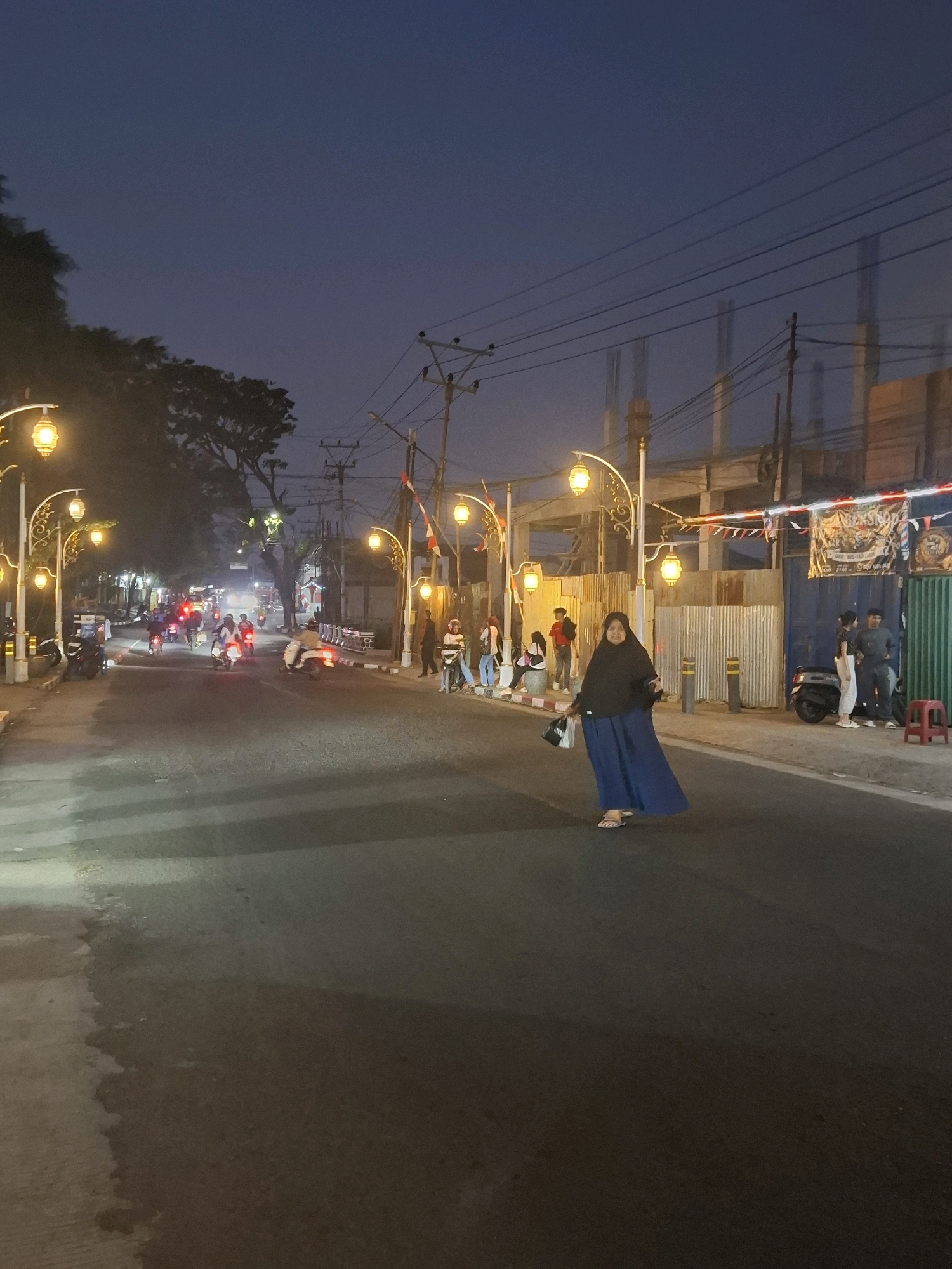 The image captures a street scene at dusk in the city of Sayang, Indonesia, with decorative streetlights illuminating the roadway. A woman wearing a black hijab and a long blue skirt is walking across the street, carrying a bag. Several people are visible in the background, some sitting and others standing near a building. Motorbikes are also present on the road, with some moving and others parked. The environment suggests a residential or commercial area, with buildings and construction in progress. The overall atmosphere is calm and illuminated by the warm glow of the streetlights against the darkening sky.