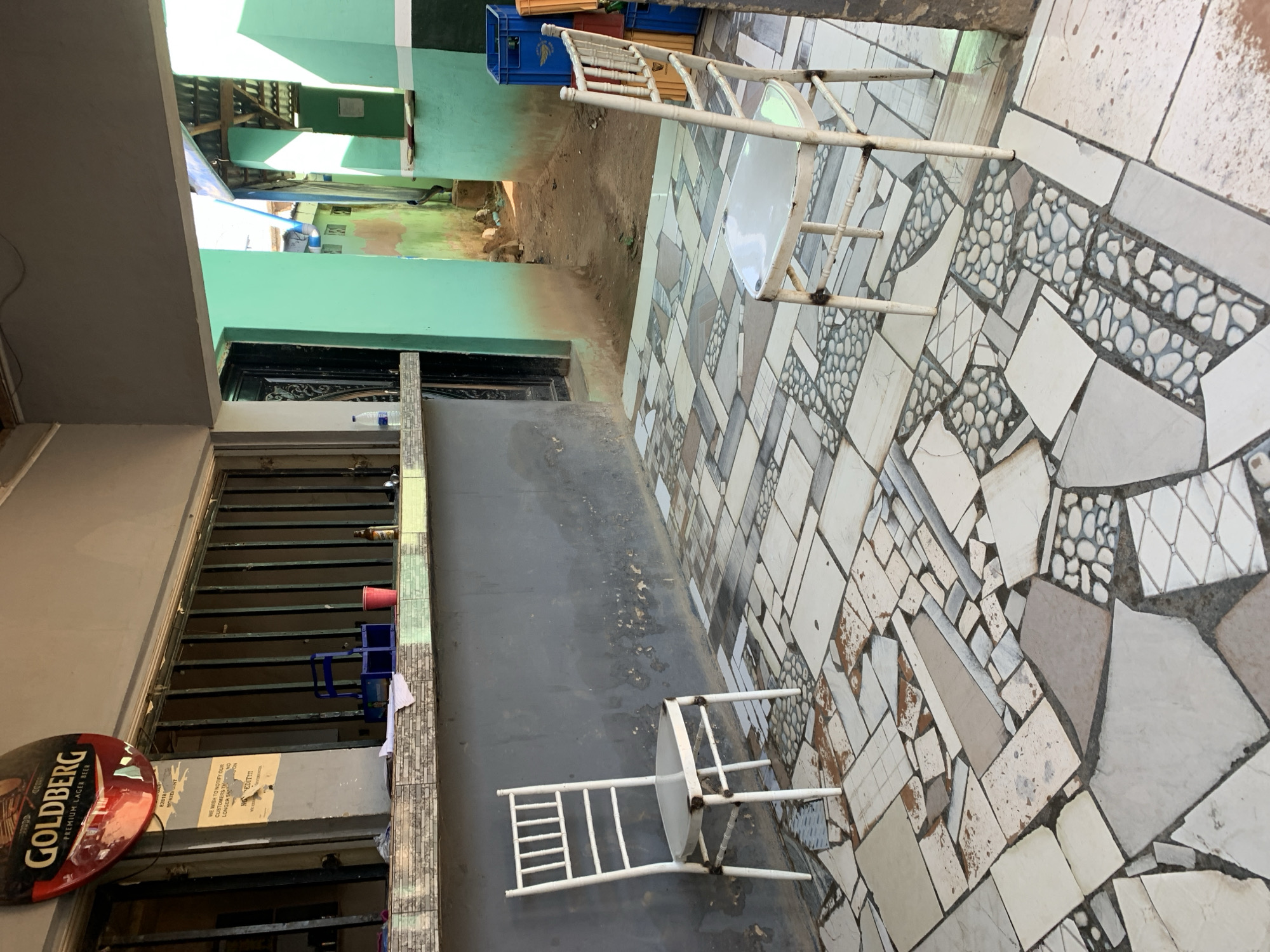 This image depicts an outdoor area, possibly a courtyard or a passage between buildings, in New Stadium, Nigeria, illuminated by bright daylight. The ground features a distinctive patchwork of various tiled patterns, including plain, textured, and pebble-inlaid sections.
In the immediate foreground, two white metal chairs are present, one standing upright and the other resting on its side on a dark grey concrete surface. To the left, a round, red, and gold sign prominently displays "GOLDBERG PREMIUM LAGER BEER," suggesting the location might be a local bar or a commercial establishment. Below the sign, a metal grate covers an opening, behind which a red plastic cup and a blue plastic container are visible on a ledge. A plastic bottle is also seen on a higher ledge.
The background shows a dirt path leading between buildings with light green-painted walls. Blue plastic crates are stacked near one of these walls. The overall scene suggests an informal, possibly commercial or semi-commercial, outdoor seating or waiting area.
Visible text includes: "GOLDBERG," "PREMIUM LAGER BEER," "82818," "MONIE," and fragments of a notice reading "WE WISH TO NOTIFY OUR CUSTOMERS TH," "STNO," "LONGER SE," and "EDIT!!!".