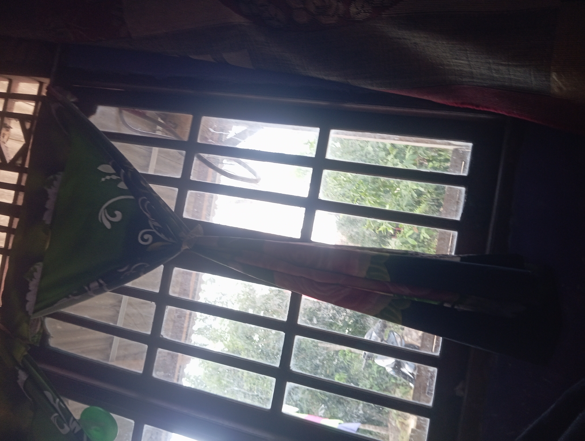 The image depicts an interior view looking out of a window. The window has a wooden frame with multiple panes, suggesting it is a casement or double-hung window. The panes are gridded by vertical and horizontal wooden bars. Through the window, lush green foliage is visible, indicating an outdoor environment with trees and vegetation. The sky appears overcast, with soft, diffused light.
Draped over the top and sides of the window are colorful curtains with a green base and white floral patterns. One curtain is partially pulled aside, revealing more of the window and the scene outside. In the lower left quadrant of the window, a portion of a motorcycle is visible, suggesting it is parked outside the building.
The lighting in the room is dim, with the light from the window being the primary illumination. The foreground of the image is dark, with hints of fabric or furniture. The overall impression is a snapshot from inside a room, looking out into a natural, somewhat rural setting.
There is no specific information in the image that allows for the determination of its location within the city of Dente Teladas, Indonesia. The scene is generic enough to be found in many locations with similar architectural styles and outdoor environments.