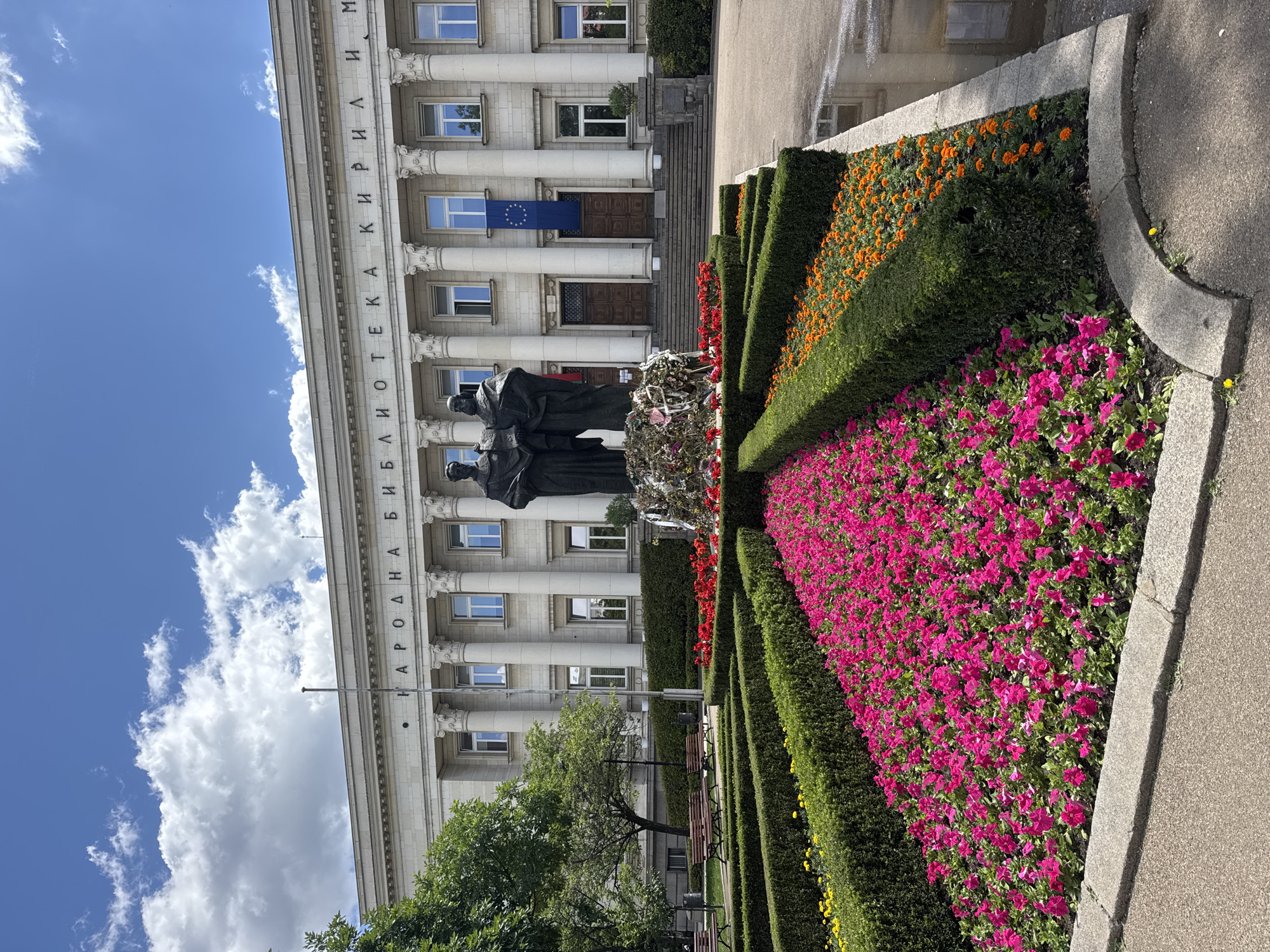 This image shows the National Library "St. St. Cyril and Methodius" in Sofia, Bulgaria. The facade of the building is visible in the background, with an EU flag hanging from it. In front of the library stands a monument dedicated to Cyril and Methodius covered in wreaths and flowers. The foreground shows a manicured flower bed with pink, orange, red, and yellow flowers bordered by low hedges. The sky is blue with some white clouds.
