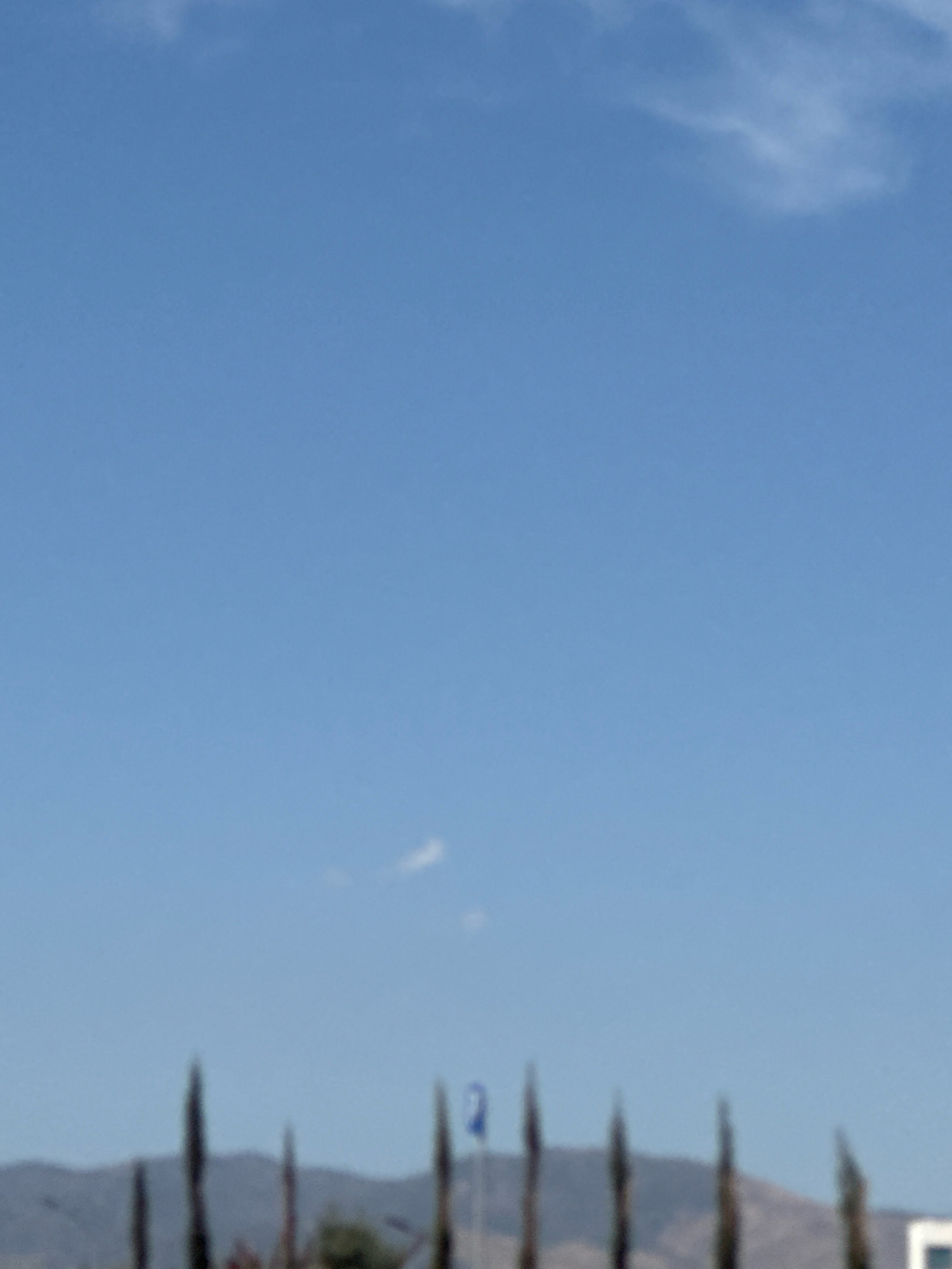 The image depicts a clear blue sky with scattered wispy clouds. In the lower portion of the frame, a line of tall, slender trees, possibly cypress, are silhouetted against a hazy background of hills. A blue flag with a white symbol, possibly a number or letter, is visible on a flagpole amidst the trees. The overall scene suggests an outdoor environment, likely during daylight hours, with a mountainous landscape in the distance. The blurry nature of the foreground elements and the distant background indicates that the photograph may have been taken from a distance or with a shallow depth of field. The location is identified as Germasogeia Municipality, Cyprus.