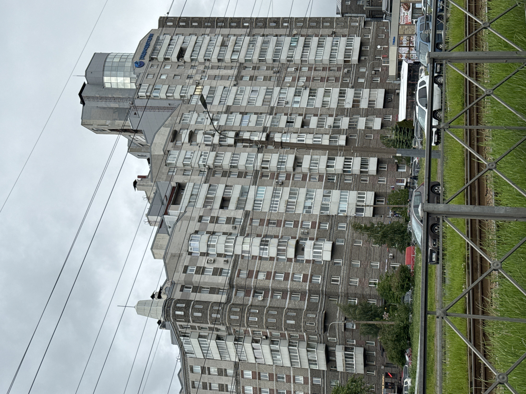 The image shows a tall, gray, multi-story residential building in Saint Petersburg, Russia. The architecture appears to be Soviet-era. The building has numerous windows and protruding balconies. Overhead power lines intersect the view of the building. In the foreground, a metal fence runs alongside what appears to be a railway track overgrown with green grass. Parked cars are visible on the right side of the image. The sky is overcast and gray. A sign on the top of the building in Cyrillic script with a logo indicates the name of the property.