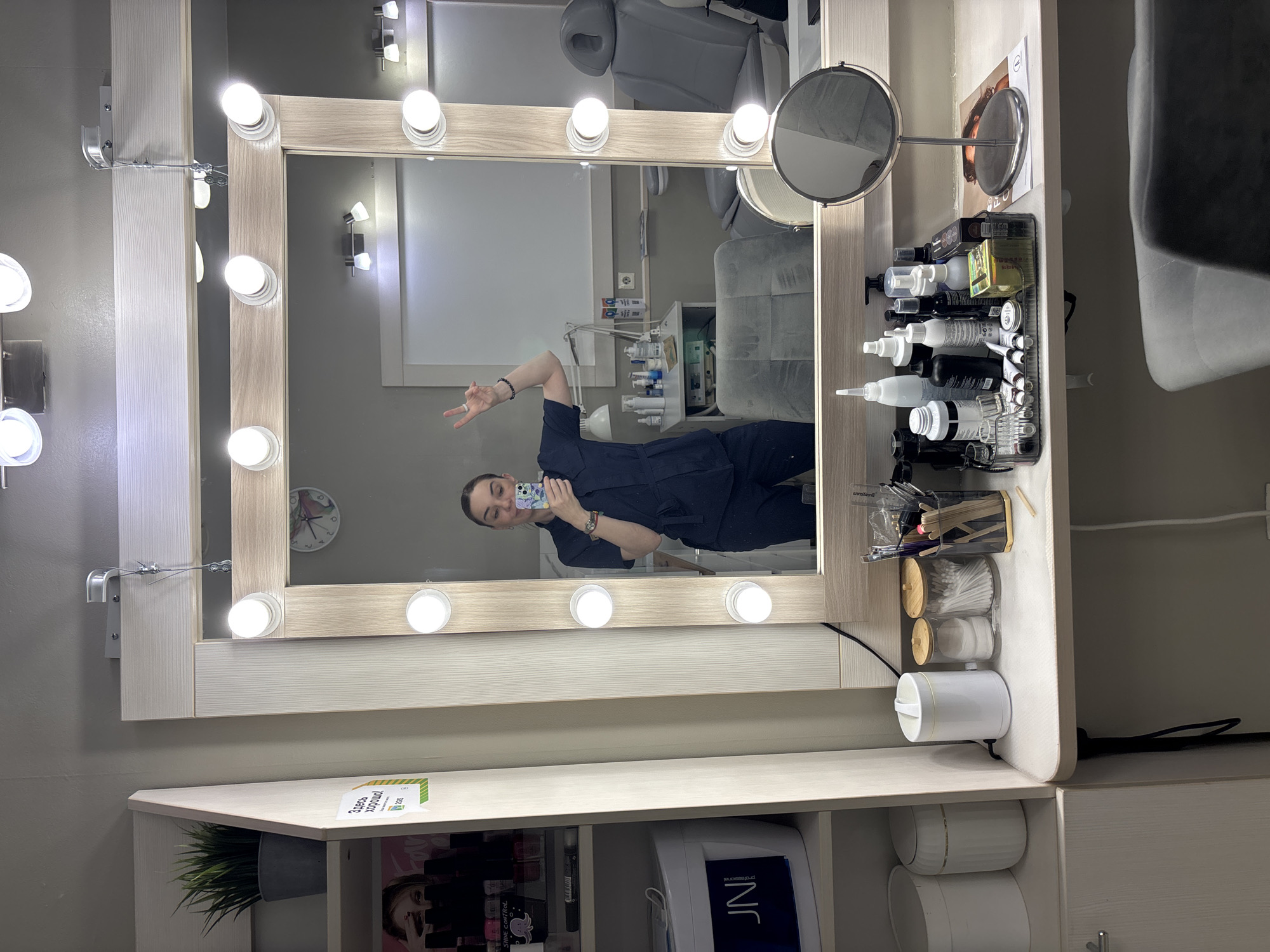 An indoor scene depicts a person taking a mirror selfie in what appears to be a beauty salon or treatment room. The image is oriented vertically, showing a large vanity mirror with a light wooden frame and multiple bright, circular bulbs illuminating its perimeter.
Reflected in the mirror is a person, likely a woman, wearing a dark blue uniform or scrubs. She has her hair pulled back and is holding a smartphone with a floral-patterned case in her right hand to capture the photo. Her left hand is raised, making a peace sign, and she has a subtle, friendly expression. The reflection shows an interior with a light grey reclining chair or treatment bed, and various pieces of salon equipment and shelving in the background. A wall clock is visible in the upper left of the reflection.
The vanity mirror is positioned above a light wooden counter that serves as a dressing table or workstation. On the right side of the counter, a black tray holds numerous bottles of beauty products, some with pump dispensers and droppers. A small, round magnifying mirror on a stand is also present. On the left side of the counter, a small artificial plant in a grey pot is visible next to a small sign.
Below the counter, there are open shelves. On the bottom left, a magazine or book featuring a person's face is partially visible. Next to it, a white rectangular device with "JNI" and "professional" printed on it can be seen, suggesting a beauty-related appliance. To the right, various beauty tools are stored in clear hanging containers, including cotton swabs and wooden sticks. Below these are white cylindrical containers.
Visible text includes "Здесь хорошо" (meaning "It's good here") and "2ГИС" on a sign on the counter. The brand "JNI professional" is on a white device. A product bottle has the barely readable text "Shine Control". The overall setting suggests a professional beauty or personal care environment.