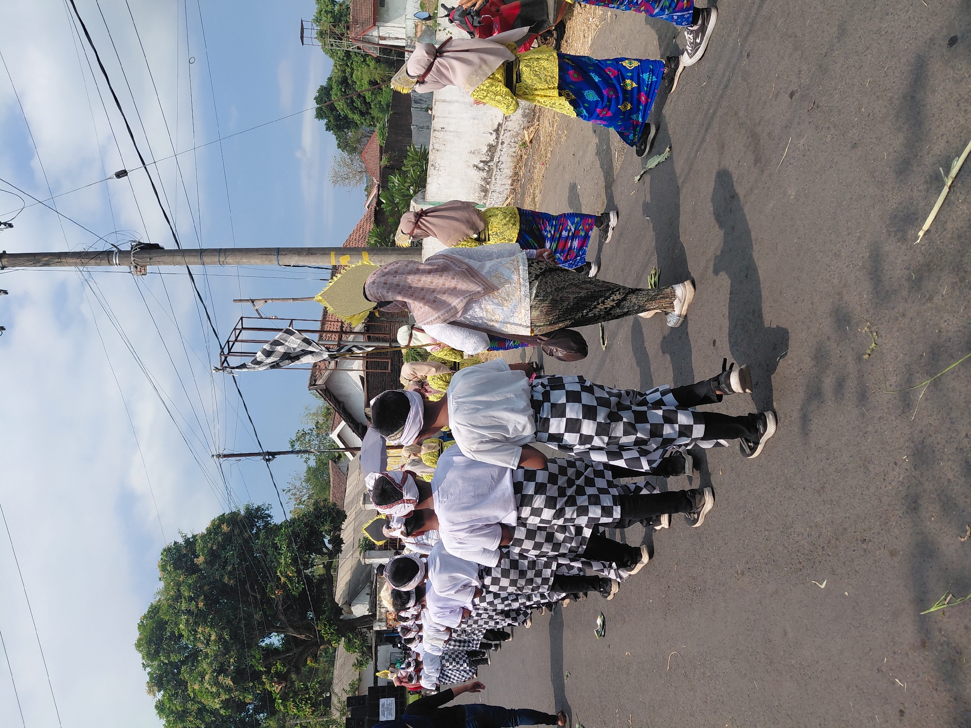 This is a street-level, eye-level, medium shot photograph of a procession or parade taking place outdoors on a paved road. The sun is shining, casting shadows on the road, and the sky is partly cloudy. The setting appears to be a village or town in Bimo, Indonesia, with residential buildings and vegetation visible in the background, including a large tree and utility poles with numerous overhead wires.
In the foreground and midground, a line of individuals, predominantly young males, are walking in procession. They are dressed in traditional attire. The males are wearing white t-shirts, black pants, and black and white checkered sarongs or kilts. They also have white headbands. Behind them, some adult females are also participating in the procession, wearing colorful traditional clothing including patterned skirts and headscarves. One woman in the midground is wearing a beige headscarf and a patterned skirt, carrying a brown bag. Another woman further back is wearing a light brown headscarf and a bright yellow top with a blue and patterned skirt.
The procession is moving from left to right in the frame. Some decorative elements, possibly part of the parade, are visible, including a black and white checkered flag on a pole and a large, golden fan-like object carried by one of the participants. Scattered on the road are dried leaves and some green stalks, suggesting a festive or ceremonial occasion. A red motorcycle is parked on the right side of the road, partially visible. On the left side of the frame, a person wearing a black top and blue jeans is walking away from the camera. A large speaker system is also visible on the far left, suggesting a musical component to the event.