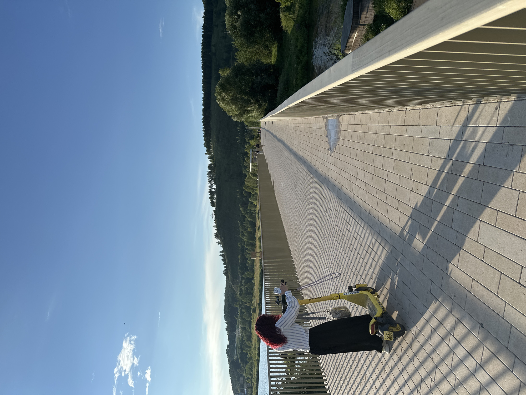 In Almetyevsk, Russia, on a sunny day, a woman with red curly hair stands on a pedestrian bridge, holding onto a yellow electric scooter. She is wearing a striped shirt and a long black skirt. The bridge has a light-colored paved surface and a light-colored metal railing. The scooter has a license plate with the numbers "0098 6832". In the background, there is a body of water, a lush green hillside covered in trees, and a clear blue sky with a few scattered clouds. The sun casts long shadows across the bridge. Further down the bridge, two figures are visible walking.