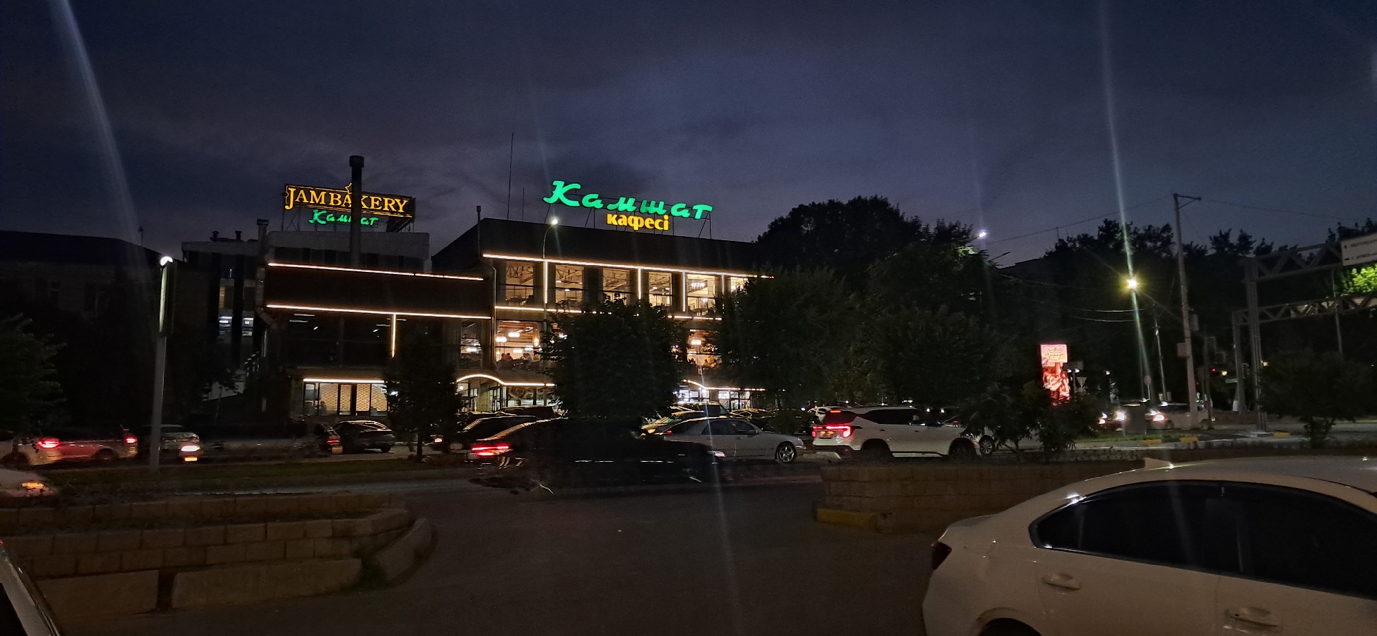 An exterior, eye-level, medium shot captures a street scene at night in an urban environment, likely in a location identified as Unknown, Unknown. The focal point is a multi-story building with illuminated signage. The top of the building displays two prominent signs: one in yellow reads "JAMBAKERY," and below it, in green, is Russian text that appears to be "Камшат кафесі" (Kamshat Cafe). The building itself features large windows on the upper floors, with interior lights on, revealing silhouettes of people inside. Decorative horizontal lights line the building's facade.
In the foreground and middle ground, several cars are visible parked and driving on the street. A white SUV is parked prominently in the middle ground, facing towards the right. Other cars, including a dark sedan with illuminated taillights and a red sedan, are also present. Trees and streetlights are scattered throughout the scene, contributing to the nighttime ambiance. The sky is dark and cloudy, typical of an evening or night setting. The overall impression is a lively commercial area illuminated by artificial lights against the dark sky.
