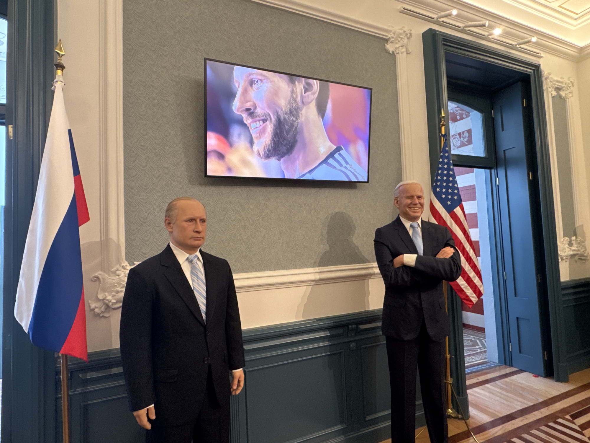 The image shows wax figures of Vladimir Putin and Joe Biden standing indoors with the flags of Russia and the United States, respectively. A television screen displays a man with a beard. The setting appears to be a room with ornate molding and a doorway. The location is the Museo de Cera in Mexico City, Mexico.