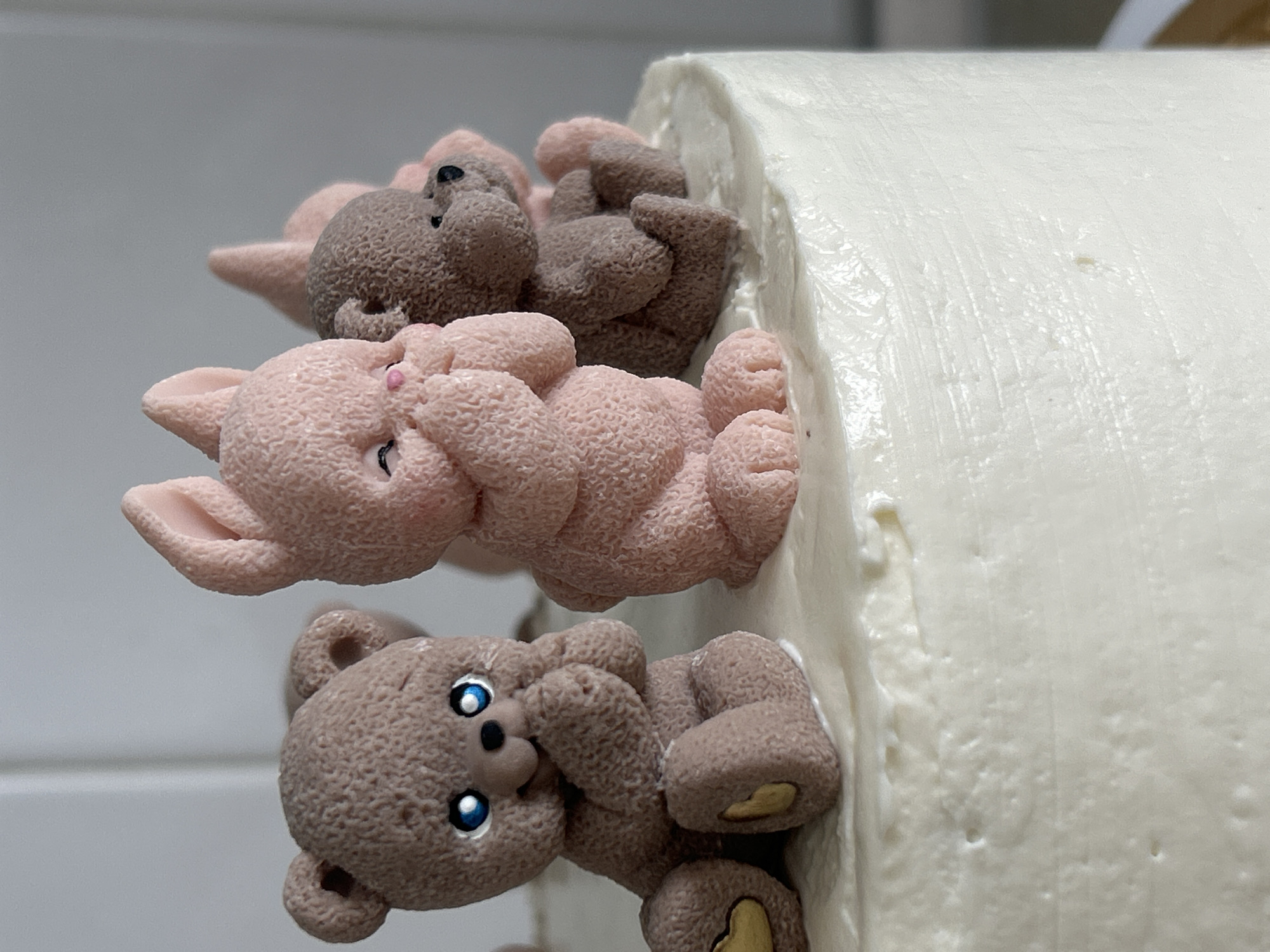 A close-up shot depicts a portion of a cake, adorned with fondant or modeling chocolate figures resembling cartoon animals, potentially bears and rabbits, in a vertical arrangement. The cake is covered in a thick layer of white frosting, appearing smooth and dense. The figures are attached to the side of the cake, one above the other, in a row. The texture of the figures suggests a matte finish, with a granulated appearance. The colors of the figures are pastel shades of brown and pink. There is no indication of the city of Chaikovsky, Russia within the scene.