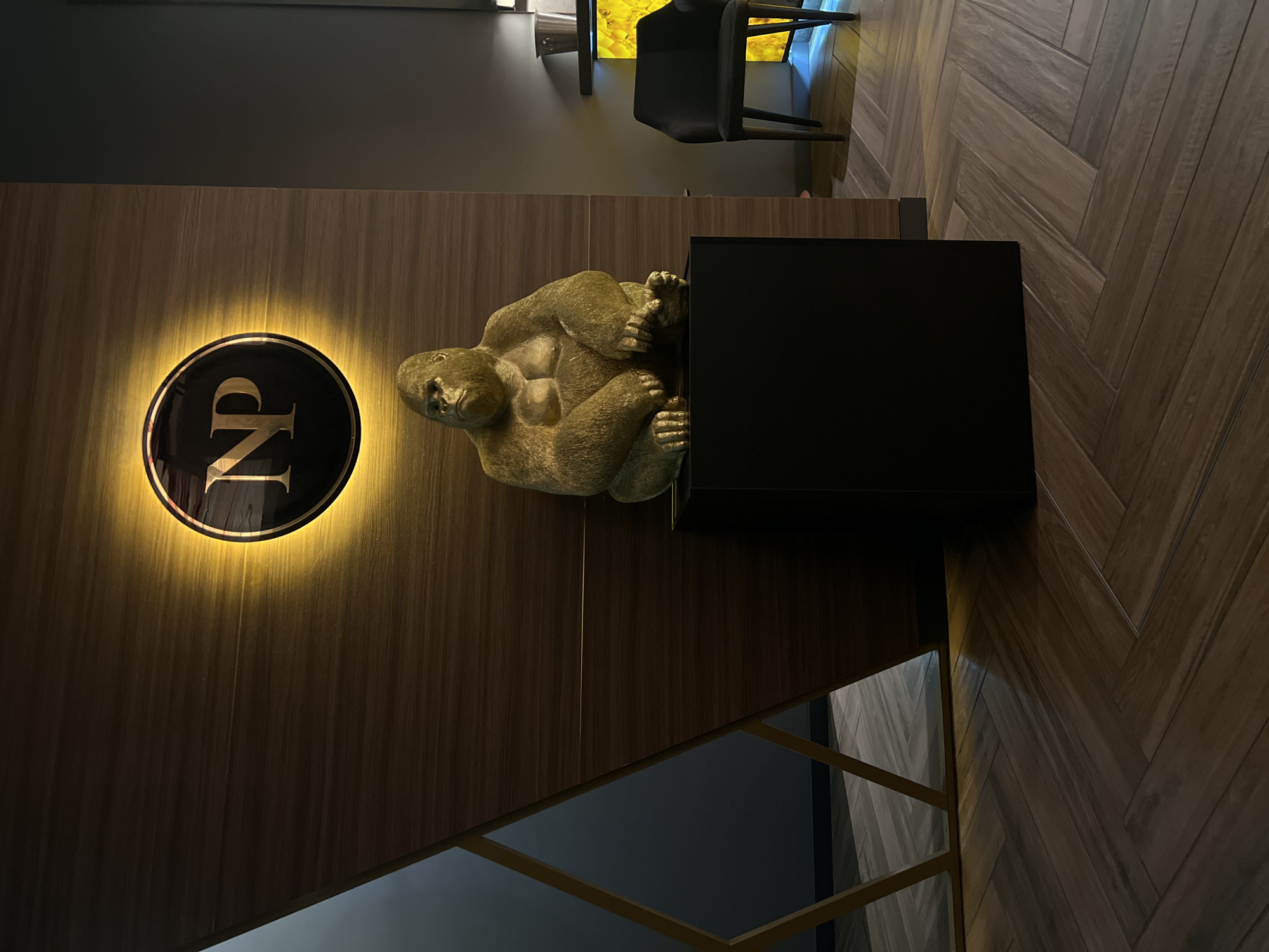 A golden gorilla statue sits on a black pedestal against a wooden wall. A circular sign with "NP" illuminated in gold is mounted on the wall above the statue. To the left, a geometric design made of gold-colored metal divides the wooden wall from a dark gray wall. To the right, a black chair sits near a shelf with a glowing yellow stone accent and a silver bucket. The floor is covered in dark wooden planks arranged in a herringbone pattern. The scene appears to be an indoor public space, possibly a lobby or lounge area, in Burgas, Bulgaria.