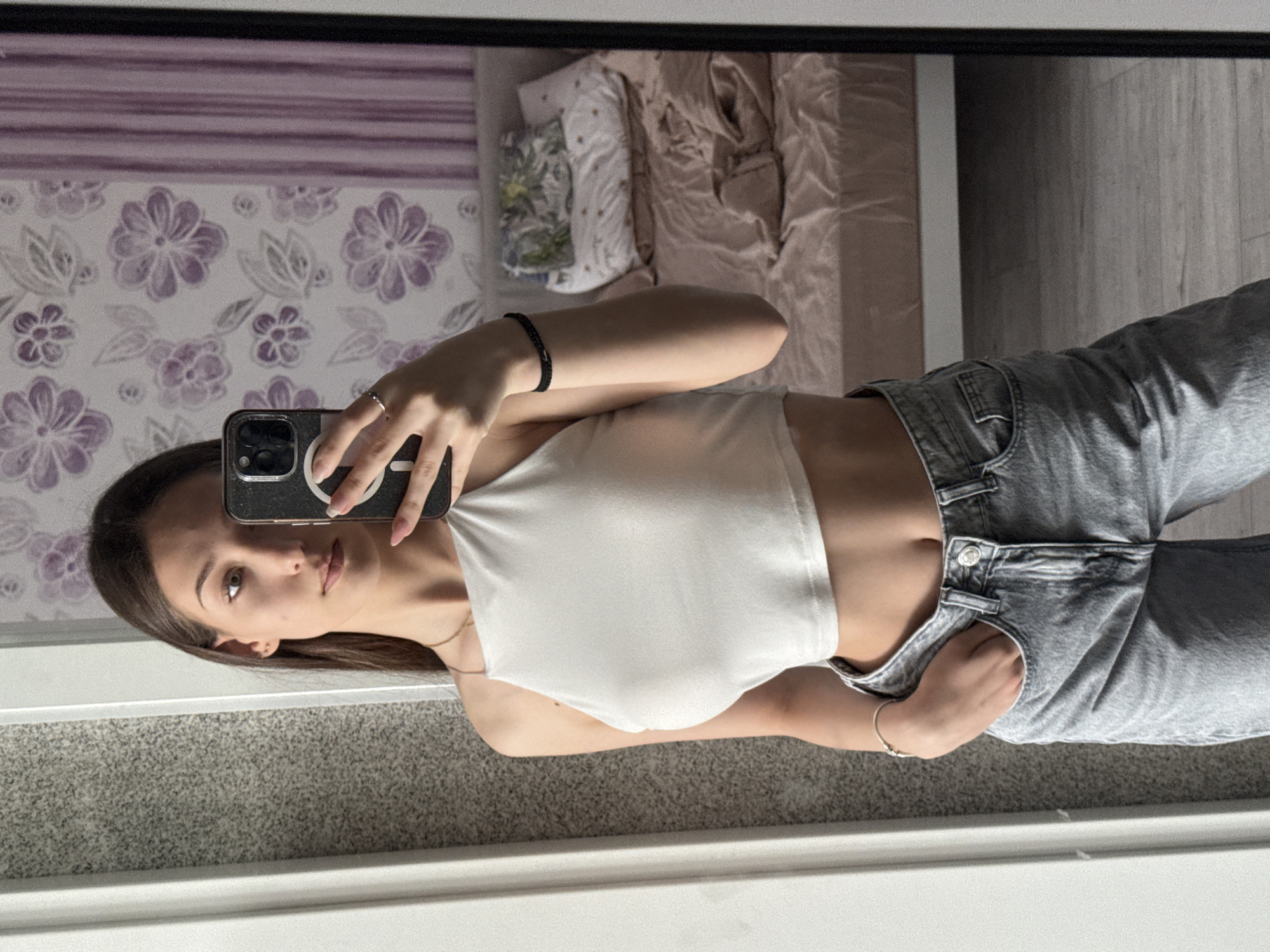 A young woman is taking a selfie in a mirror. She is wearing a white halter top and gray jeans. Her left hand is holding a phone, and her right hand is in her pocket. She has a bracelet on her right wrist and a ring on the fingers of her left hand. The background includes floral wallpaper, bedding, and carpeting. It is not possible to ascertain the location of Sofia, Bulgaria from the image.