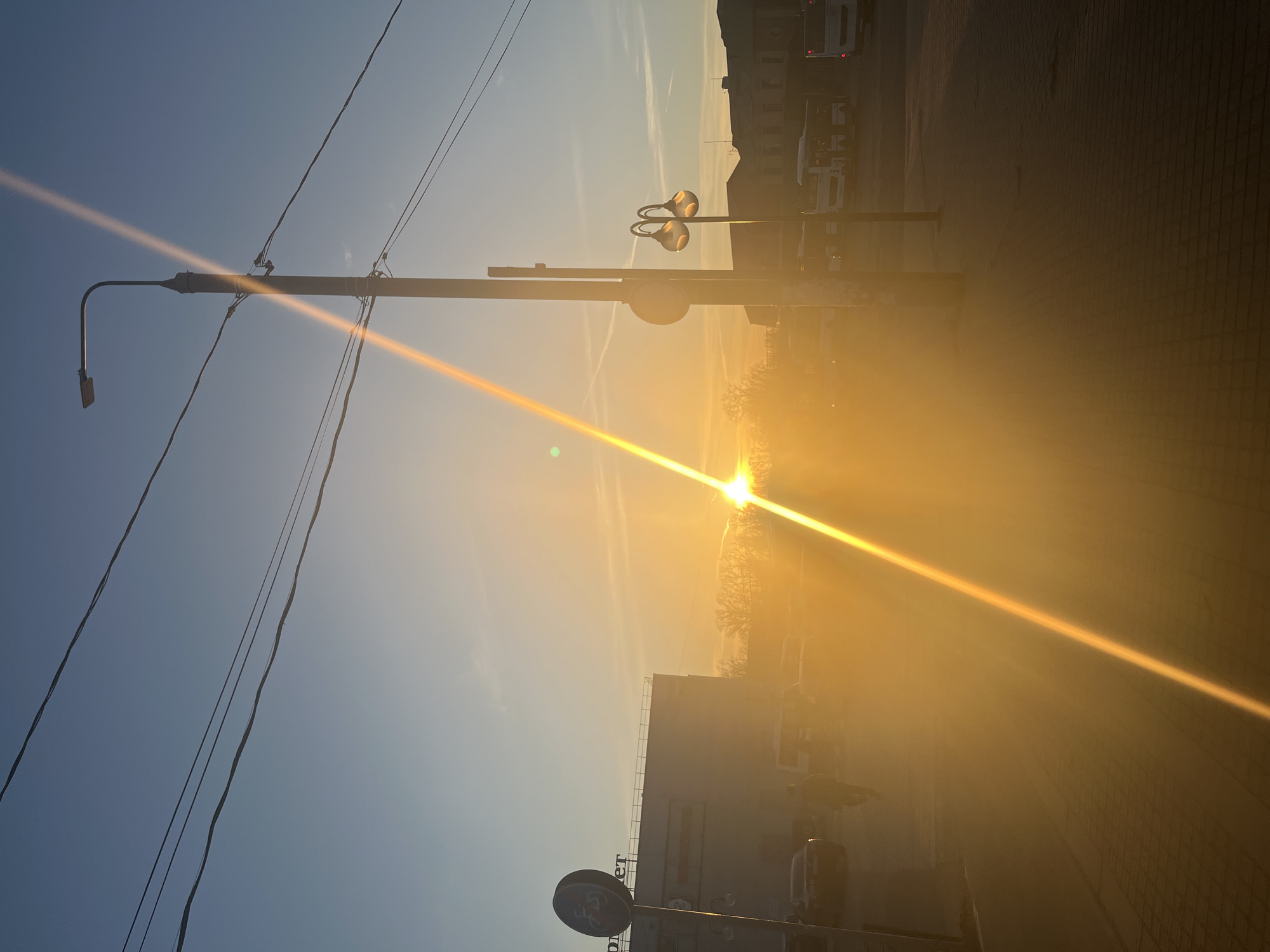 The image displays an urban street scene at Privokzalnaya Square, Klin, Russia, adjacent to the "Orion" Shopping Center, under low sun conditions, likely sunrise or sunset. A strong golden lens flare originates from the sun positioned in the mid-left of the frame, diagonally traversing the image.
In the foreground, a dark-toned brick paver sidewalk extends. A single blurred human figure is visible walking on this sidewalk toward the left.
On the left side, a multi-story building, identified by a partially visible sign displaying "ОРИОН" and "ЛЕТ," features a light-colored facade and a flat roof. In front of this building stands a circular regulatory sign, black with a white border, depicting a white bicycle with a red diagonal line through it (No Bicycles). Several indistinct vehicles, including a dark sedan, are present in the area before the building.
The right side of the image features two distinct lampposts. The taller, left-most pole is a grey utility pole equipped with a modern, rectangular LED streetlamp fixture and multiple dark utility wires. To its right and slightly shorter, a decorative lamppost with two globe-shaped luminaires extends. Behind these, a series of white passenger buses are parked or idling, indicative of a bus station. Beyond the buses, older, multi-story buildings with pitched roofs are discernible.
The background shows a line of deciduous trees without leaves, silhouetted against the bright light. The sky transitions from a brilliant golden-orange at the horizon around the sun to a clear light blue overhead, with faint white contrails visible. Multiple dark utility wires span the upper portion of the frame.