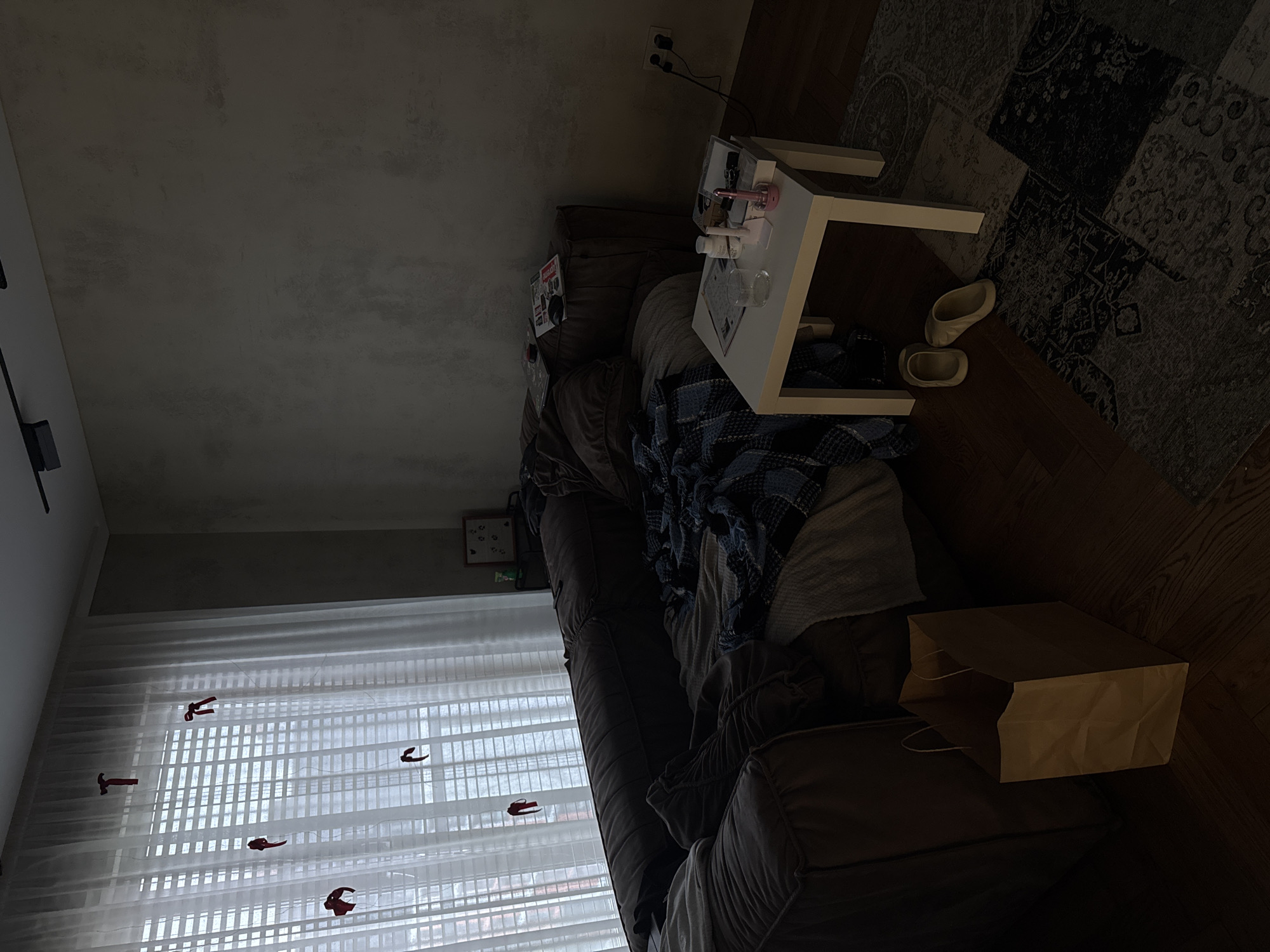 The image depicts an indoor scene, likely a living room, with dim lighting. A large brown sofa dominates the left side of the frame, partially covered with blankets and pillows, suggesting it's being used as a bed. In front of the sofa, a small white table holds a glass of water, some toiletries, and a pink electronic device. To the right of the table, a pair of light-colored slippers rests on a patchwork rug. A brown paper shopping bag is positioned to the left of the sofa. The background features a large window with vertical blinds, through which some red objects, possibly decorations, are visible hanging. The wall behind the sofa has a textured, gray finish. The overall impression is one of a lived-in space. No individuals are present in the image, and there is no explicit information to pinpoint the exact location within Бараўлянскі сельскі Савет, Belarus.