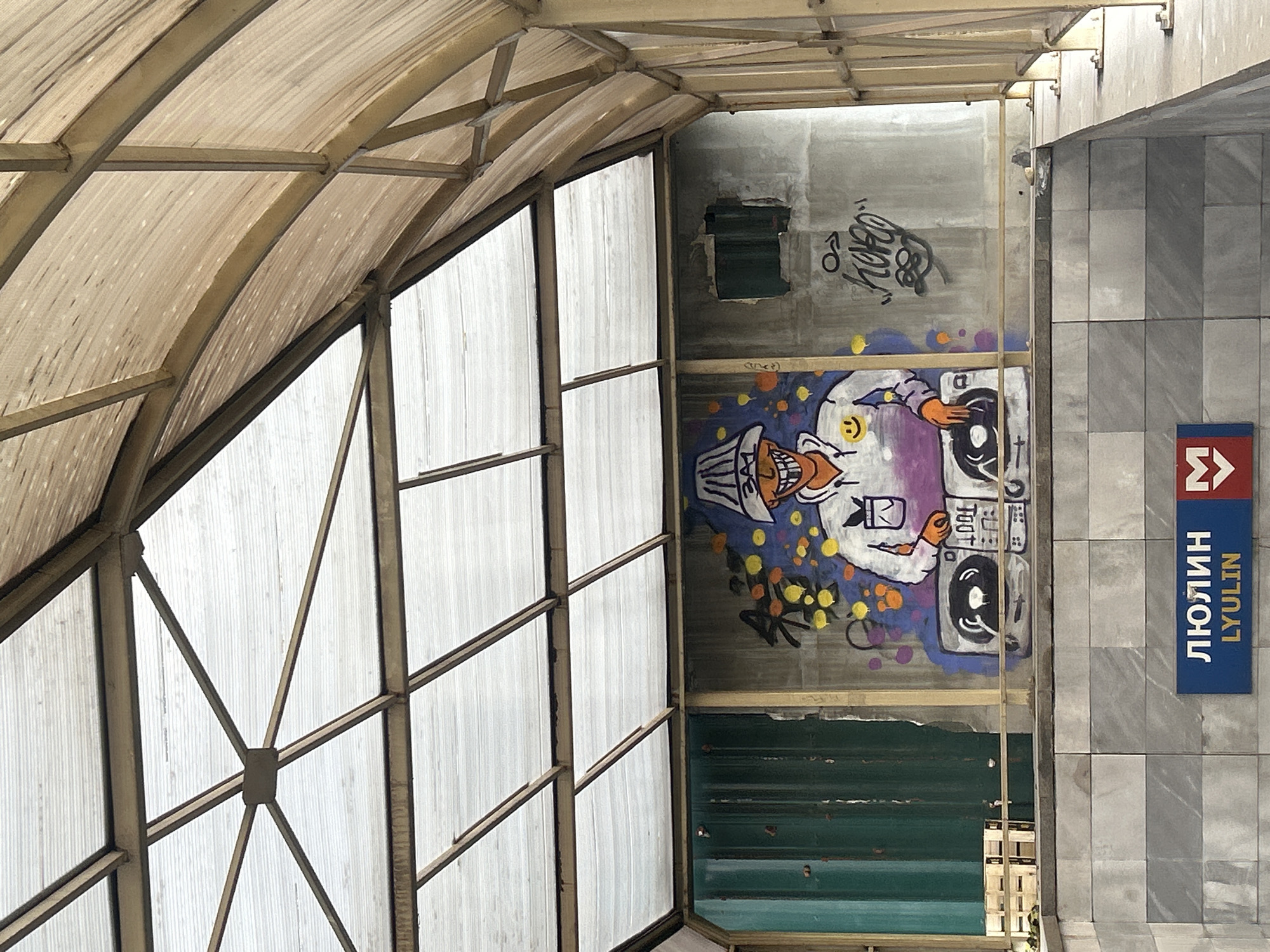 The image captures a view from below a structure, possibly a covered walkway or entrance, in the city of Unknown, Unknown. The structure consists of a metal framework covered by a semi-transparent material. To the right, a concrete wall features graffiti, including a cartoonish character and text. Next to the wall is a blue sign with white lettering in Cyrillic script and its English transliteration, "LYULIN," along with a symbol associated with public transportation. The ground is paved with large, light-gray tiles.