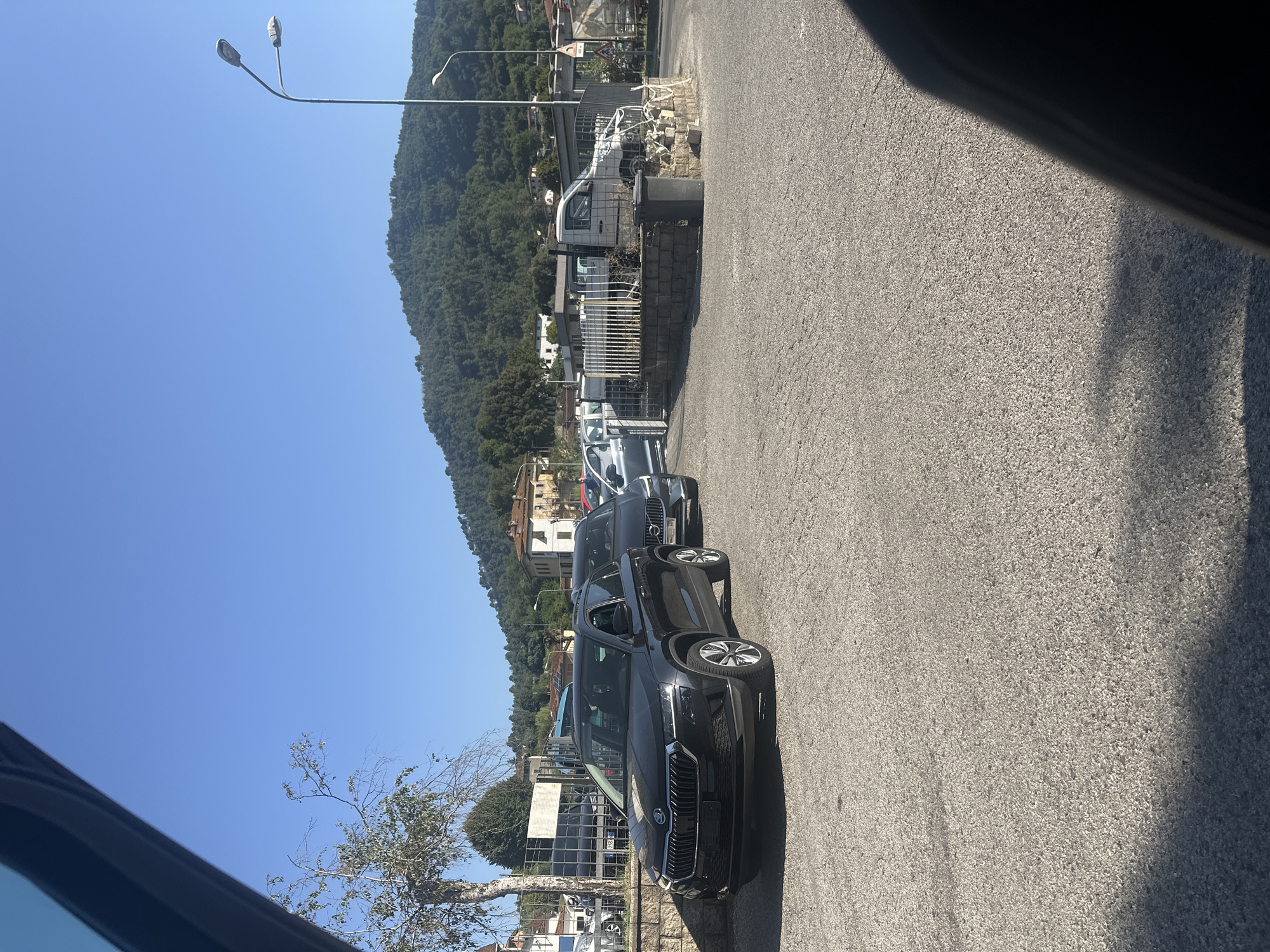 In Fornoli, Italy, a black Skoda Karoq is parked on a gravel lot under a clear blue sky. The car is positioned at an angle, facing towards the left of the frame. Behind it, another dark-colored SUV and a white utility vehicle are visible, partially obscured by a metal fence. The background features a hillside covered in trees and some buildings, suggesting a semi-urban or rural setting. A street light stands tall to the right of the scene. The perspective suggests the photograph was taken from inside another vehicle, with a portion of its interior visible in the foreground.