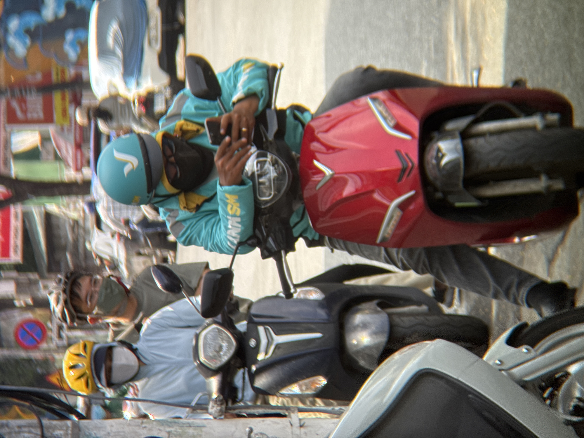 The image captures a street scene in Nha Trang, Vietnam, focusing on three individuals on motorcycles. In the foreground, a person wearing a turquoise jacket with yellow accents and a turquoise helmet with a white "V" logo is operating a red scooter and looking at a smartphone. They are wearing a dark face mask. To their left, another person on a dark scooter wears a light blue jacket, a white face mask, and a yellow helmet. Behind them, a third person on a motorcycle wears a green face mask and a darker helmet. The background shows a blurry urban environment with buildings and traffic, consistent with a city in Vietnam. The lighting suggests it is daytime.
