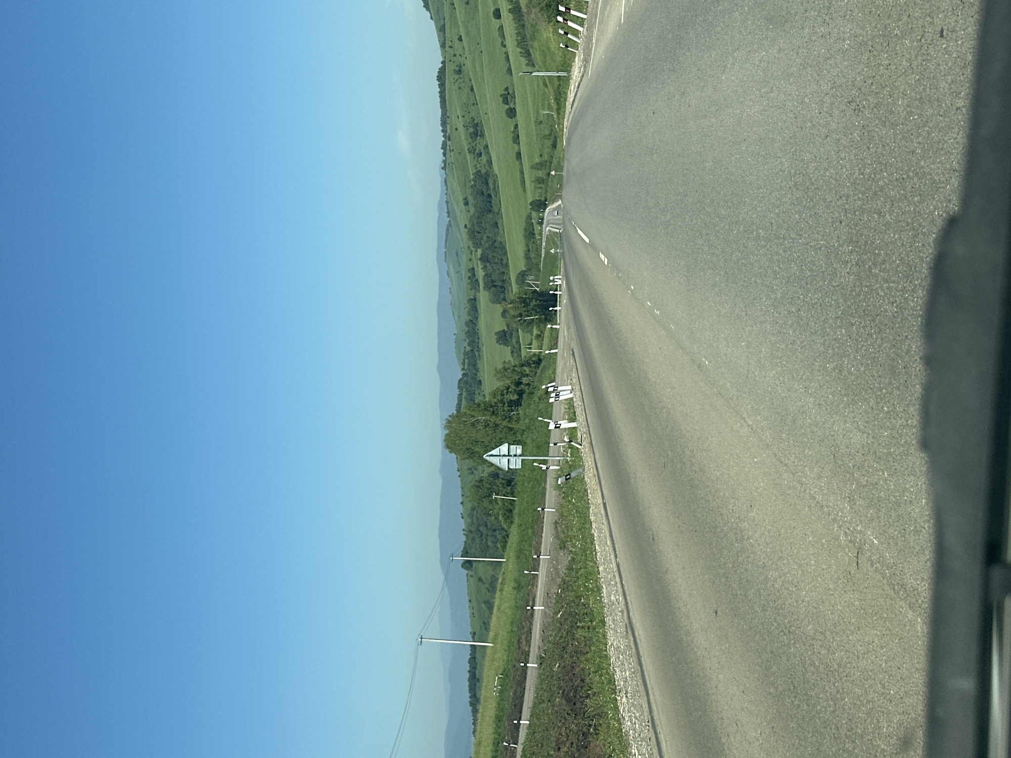 A paved road curves through rolling green hills under a clear blue sky. The road is lined with white and black markers, and on the left side, there are power lines supported by poles. In the distance, the road leads towards more hills, with a few vehicles visible. The foreground is obscured by a dark shadow, possibly from a vehicle. The scene appears to be in a rural or semi-rural area, consistent with a location like Verkh-Aya, Russia.