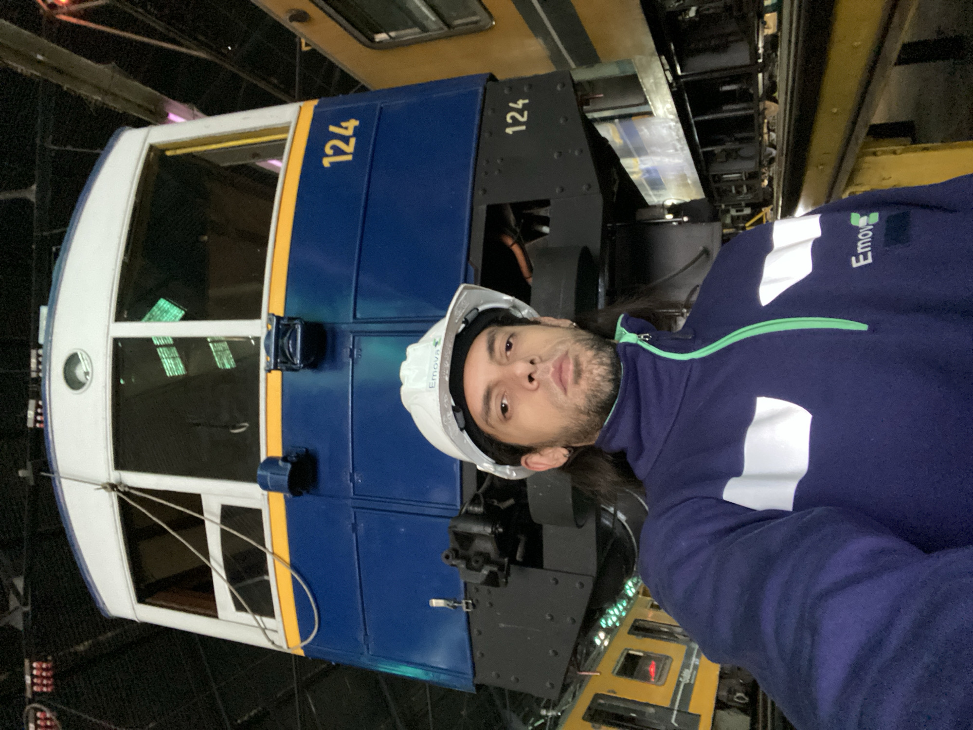The image depicts a man in a white hard hat and blue "Emova" branded work jacket standing in front of a blue and white train car with the number "124" on its side. The environment appears to be a maintenance or storage facility with other train cars visible in the background. The location is likely Buenos Aires, Argentina, based on the Emova branding, which is associated with the Buenos Aires Underground system.