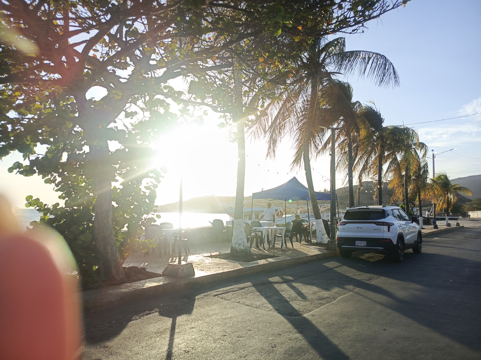 The image shows a street scene in Juan Griego, Venezuela. A white SUV is parked on the right side of the road, with its rear facing the camera. To the left, a waterfront area is visible with tables and chairs under a blue canopy, presumably a restaurant or outdoor seating area. People are sitting and standing in this area. The scene is bright, with sunlight filtering through trees, including palm trees, along the waterfront. The road is paved and shows shadows cast by the trees and the car. A blurry object obstructs a portion of the left-hand-side view.