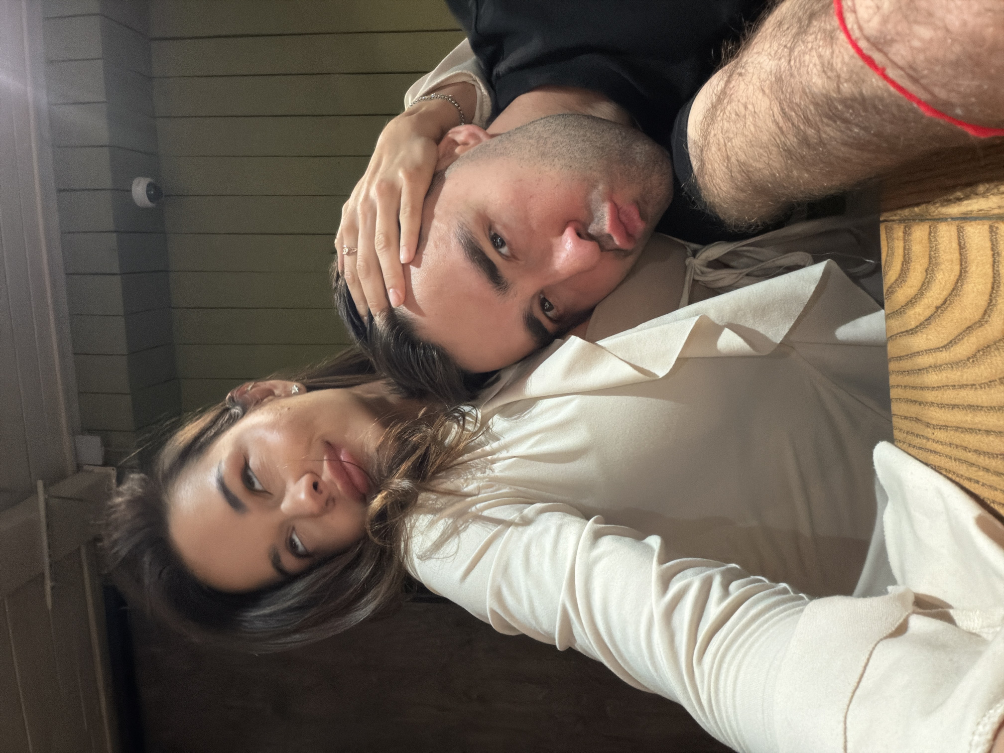 This rotated close-up selfie features a man and a woman, likely a couple, in an indoor setting.
The man, positioned in the upper section of the frame, has dark hair and a beard. He looks directly at the camera with a playful expression, sticking his tongue out slightly. He is wearing a black shirt, and his hairy right arm, adorned with a red string bracelet, is visible in the foreground. A woman's hand, wearing a silver chain bracelet and a ring with a small stone on her ring finger, rests on his head.
The woman is in the lower part of the frame, looking towards the camera with a subtle pout. She has long dark hair and is dressed in a light-colored, possibly cream or beige, long-sleeved top or dress, featuring ruffled details around the neckline or shoulder. The fabric of her clothing exhibits distinct folds and texture.
The background shows wooden plank walls painted a muted green or gray. A small white security camera is mounted on one of these planks in the upper left. A segment of a wooden table with a visible grain pattern is present in the lower right, suggesting a cafe, restaurant, or a cozy home interior in Krasnoyarsk, Russia. The lighting is characteristic of indoor artificial light. The image captures a casual and affectionate moment, highlighted by the man's playful expression. No text is visible.
