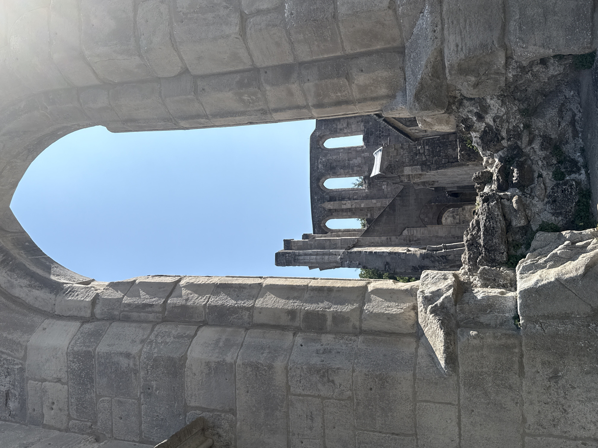 The image shows the ruins of the Walkenried Abbey in Walkenried, Germany, viewed through a large stone archway. The abbey ruins consist of weathered stone structures with arched windows and crumbling walls. Vegetation is visible growing amidst the rubble. The sky is clear and blue, suggesting a sunny day. The foreground features large, rough-hewn stone blocks, part of the structure framing the view of the ruins. The perspective is from within the abbey grounds, looking outwards towards the main surviving structures.